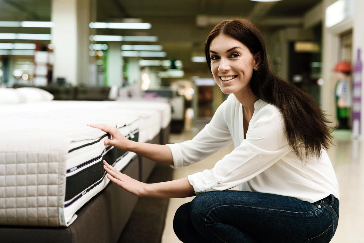 A smiling woman crouches beside a mattress in a store, pressing its side to test firmness or construction.
