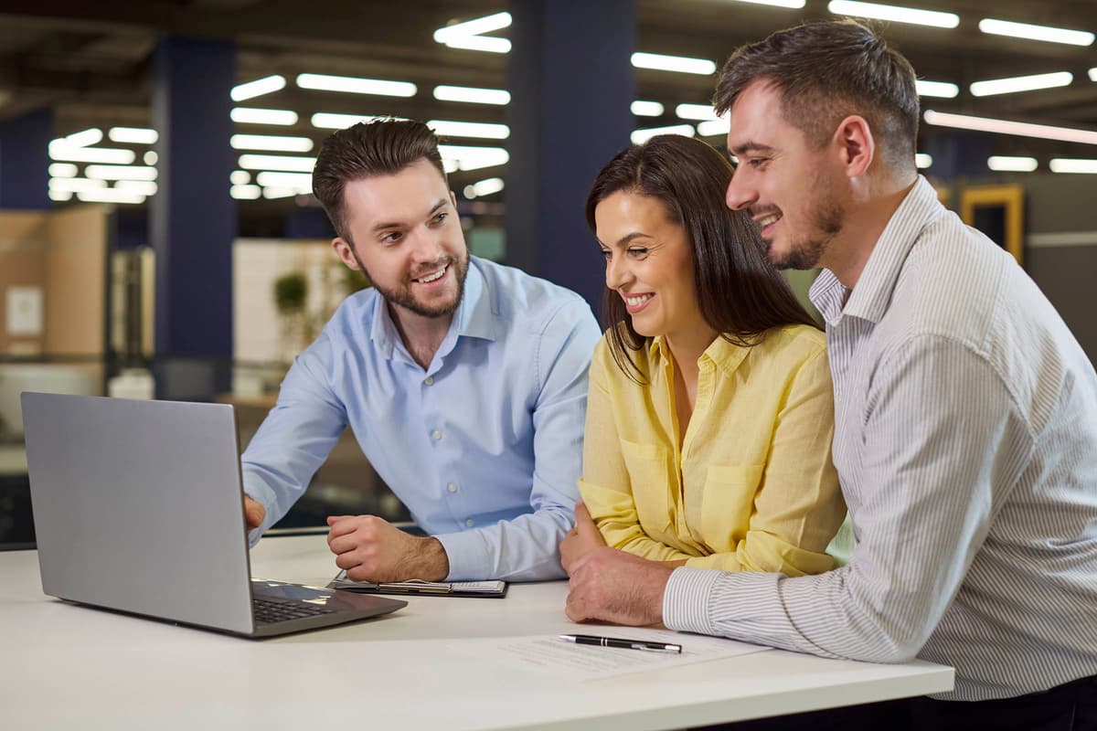 Three colleagues smiling and discussing work at a desk, looking at a laptop in a modern office environment.