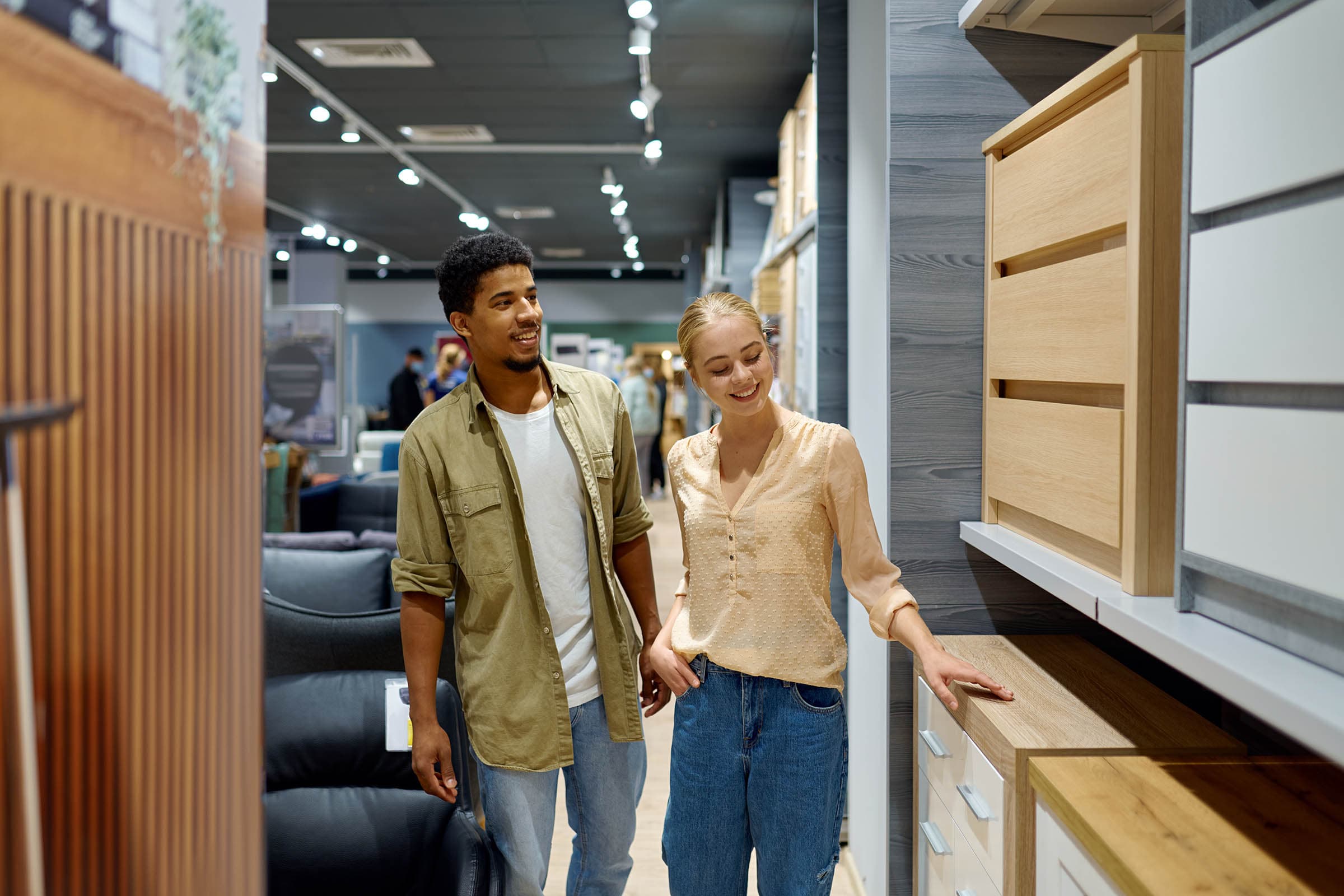 A couple shopping in a furniture store, smiling and looking at wooden cabinets displayed on shelves.