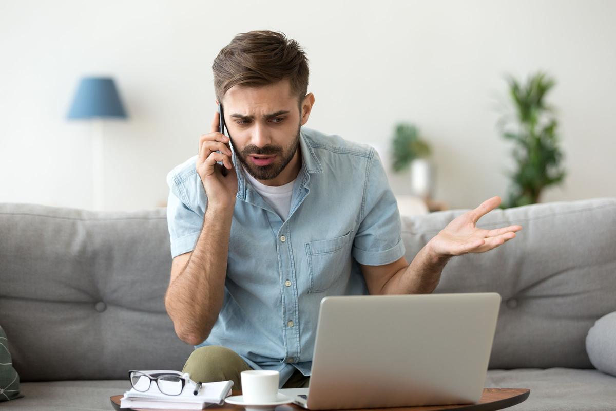 Frustrated man sitting on a couch, talking on the phone while looking at a laptop, with a notepad and coffee cup nearby.