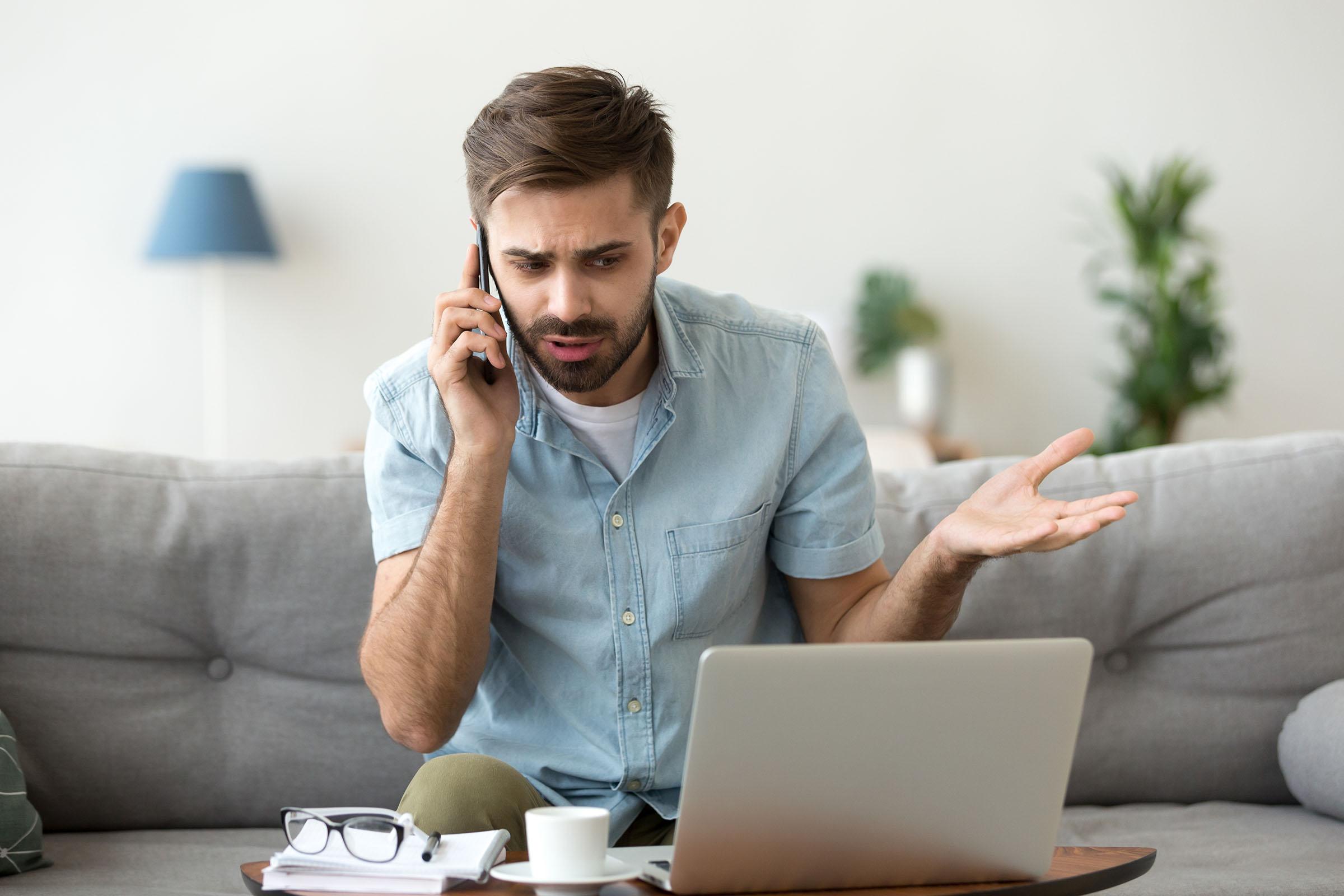 Frustrated man sitting on a couch, talking on the phone while looking at a laptop, with a notepad and coffee cup nearby.