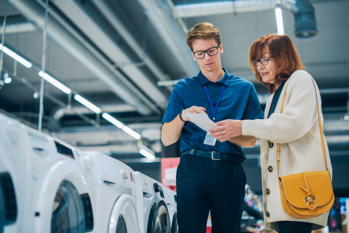 A store employee assists a woman with a purchase, examining a receipt in an appliance showroom.