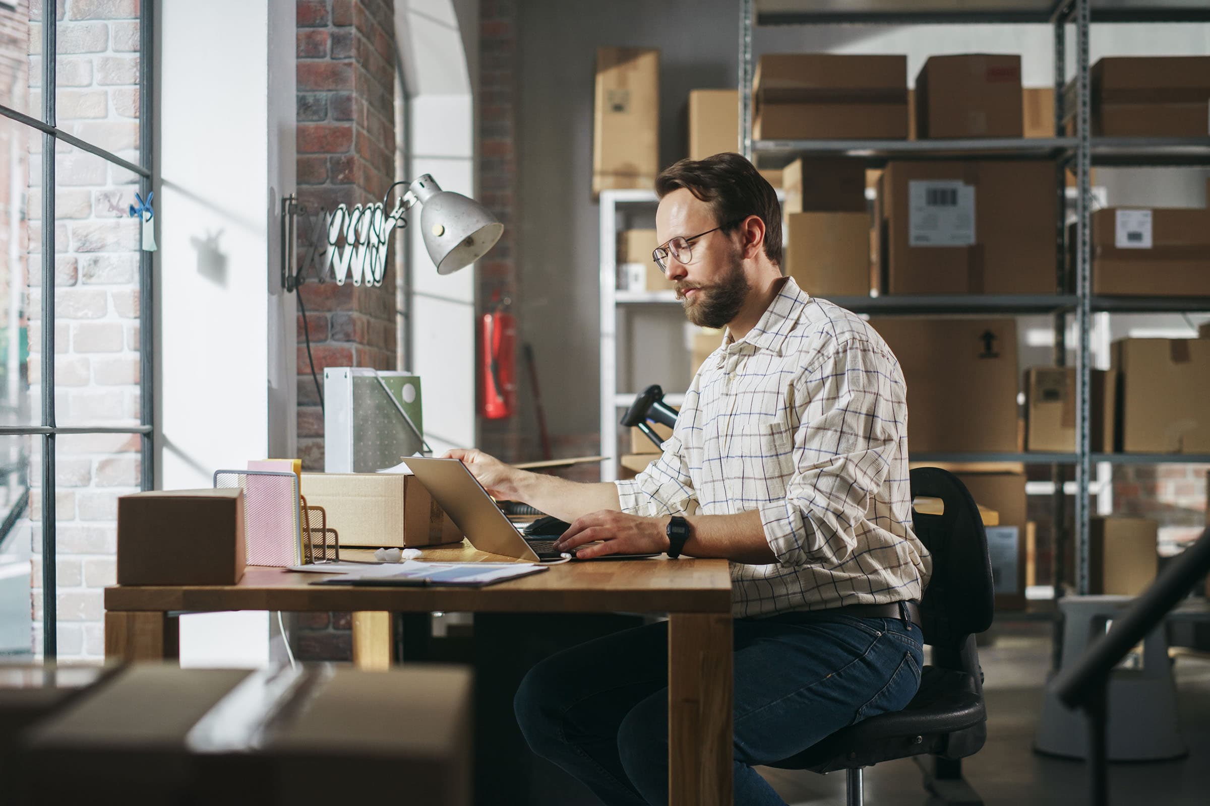 Man working at a desk with a laptop in a warehouse, surrounded by boxes and shelves, under a window with natural light.