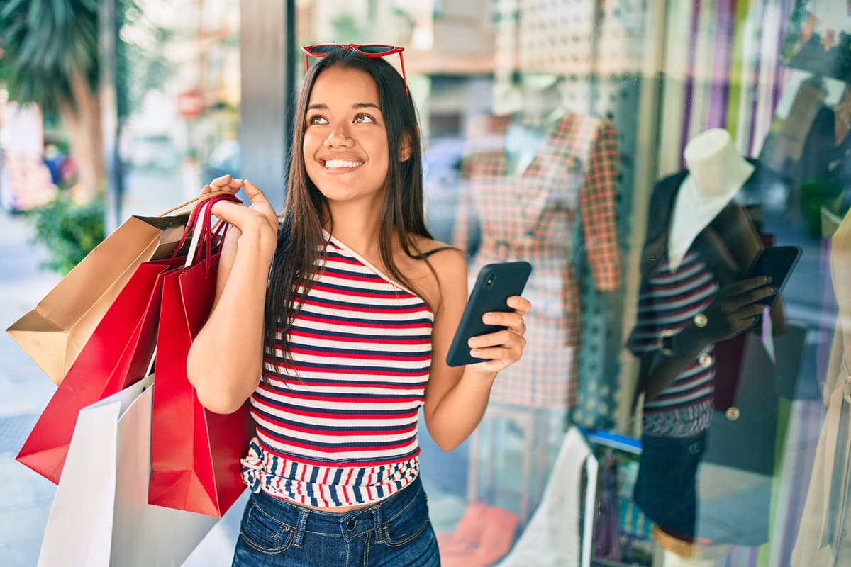Smiling woman holding shopping bags and a smartphone stands outside a clothing store window, wearing a striped top and jeans.