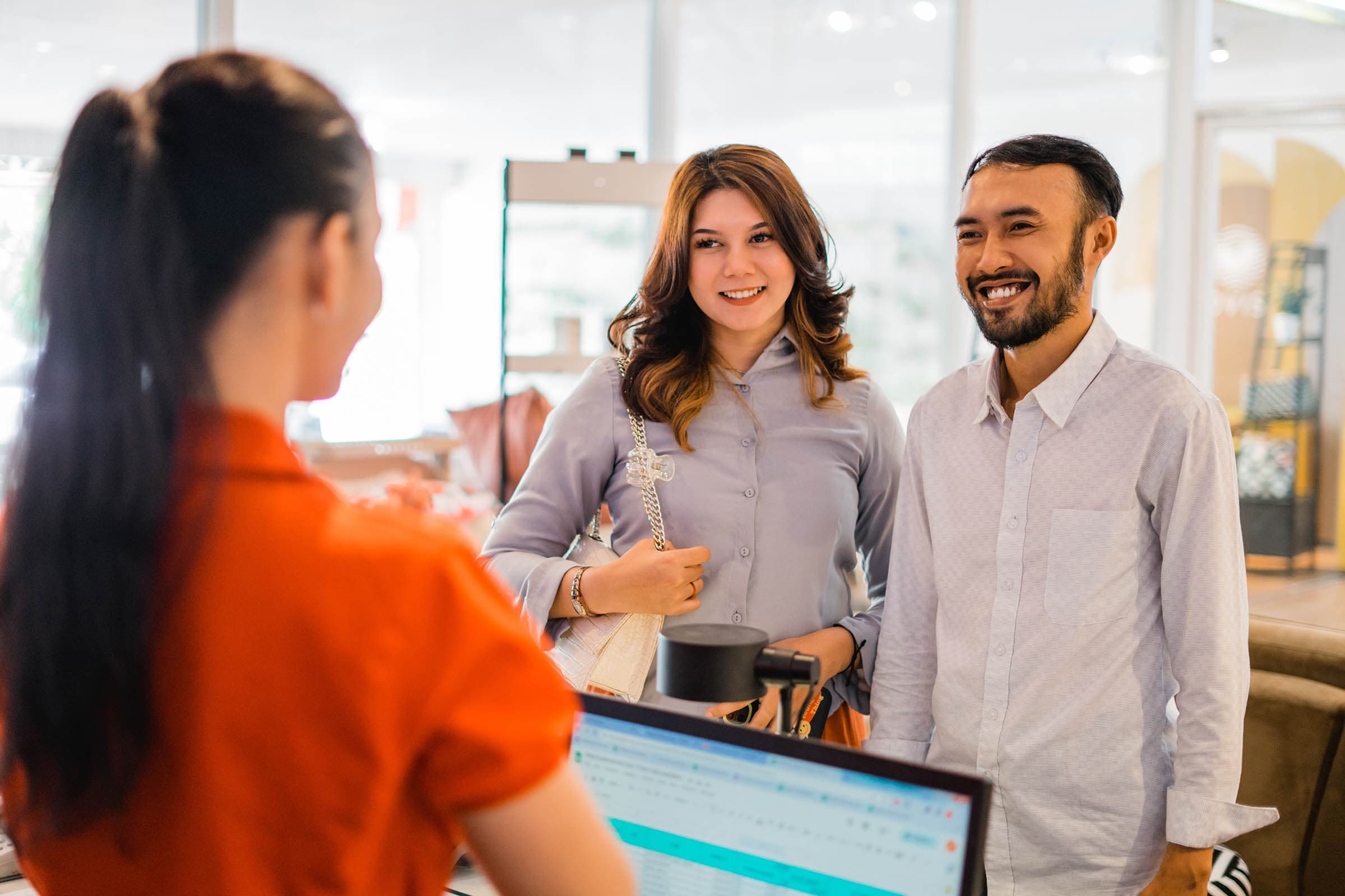 A couple smiles at a receptionist in a bright, modern office setting, viewed from behind the receptionist.
