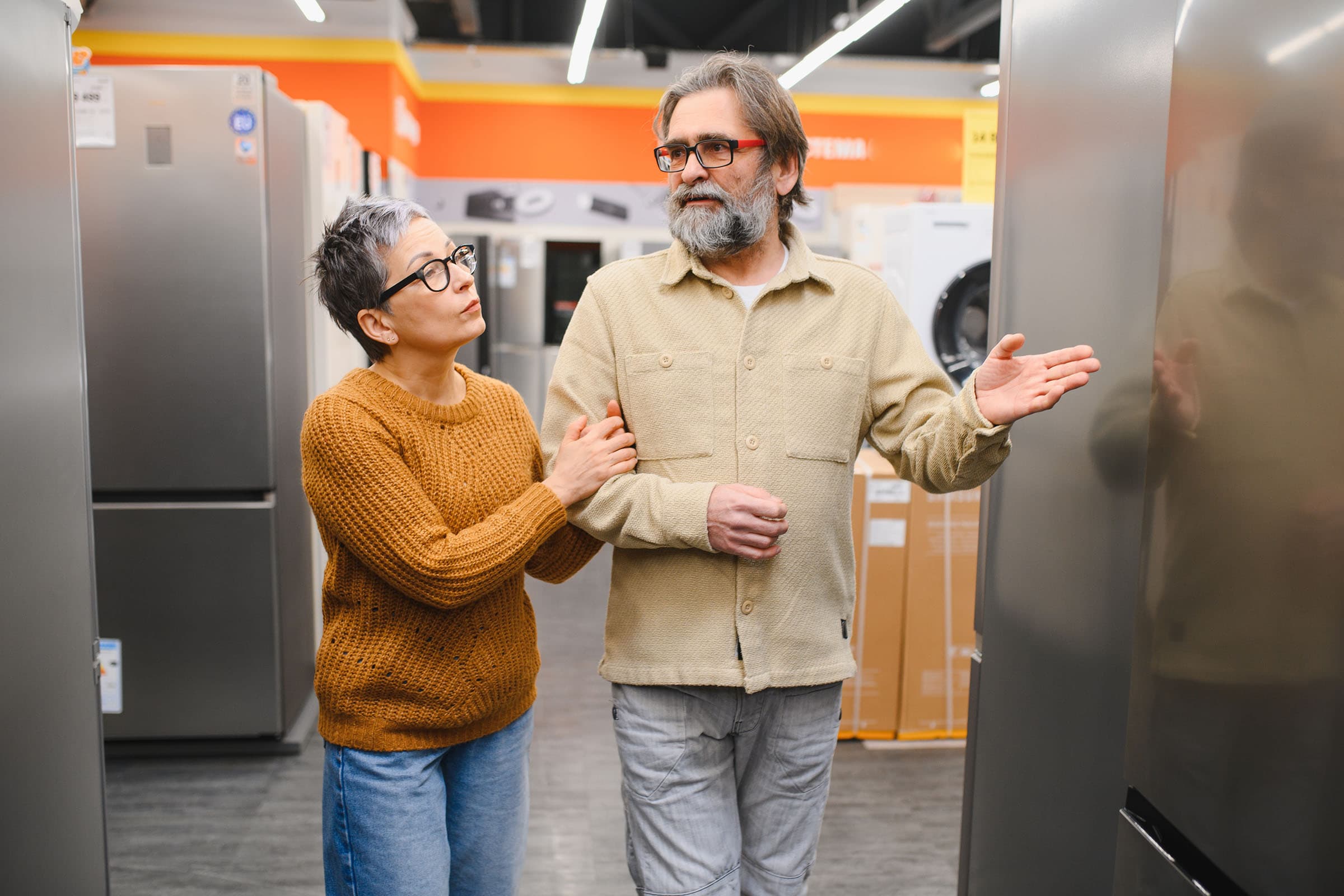 A couple, arm in arm, is discussing fridges in an appliance store, surrounded by various models and bright lighting.