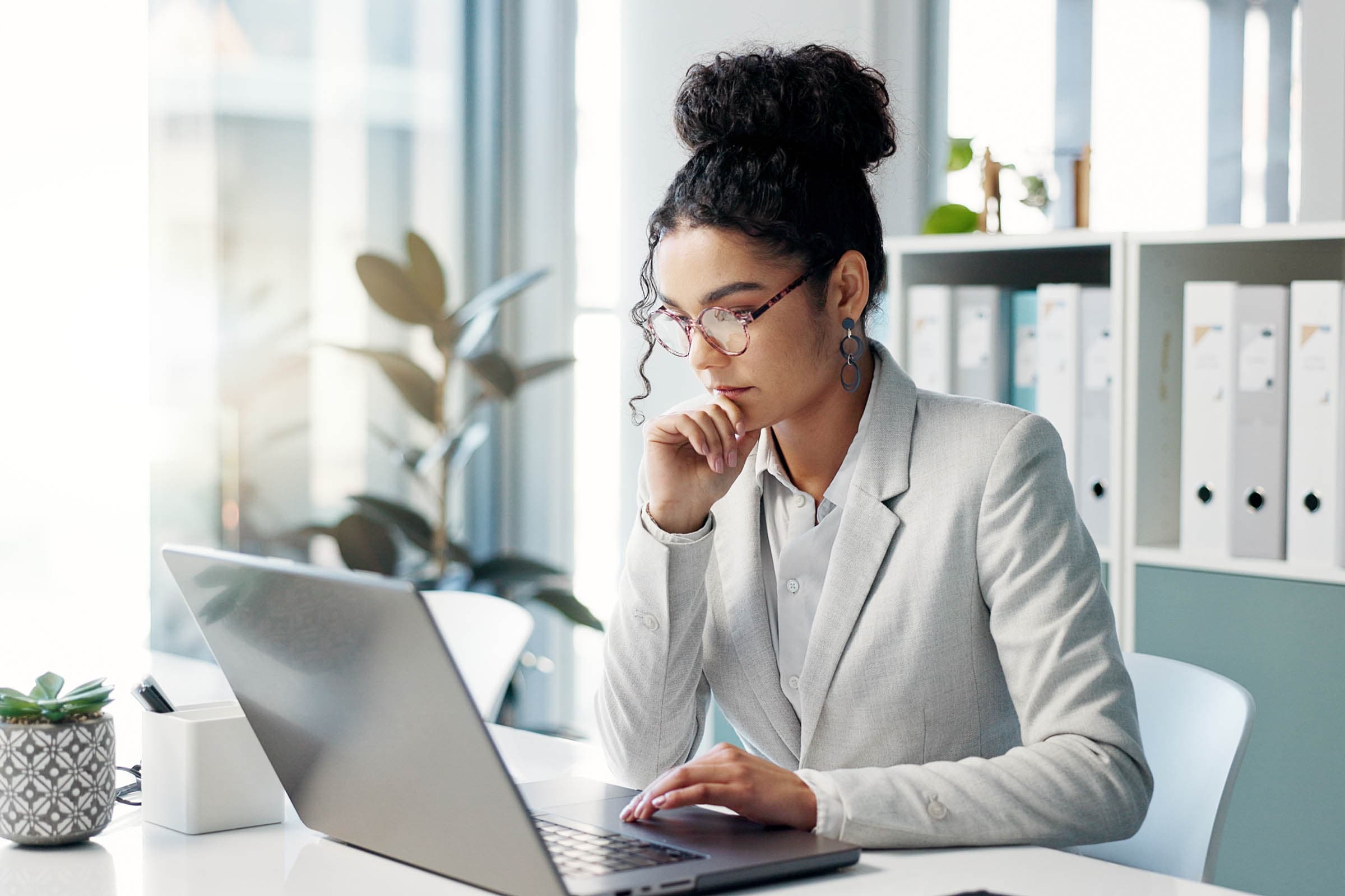 A woman in a gray suit works intently on a laptop in a bright office, with plants and shelves in the background.