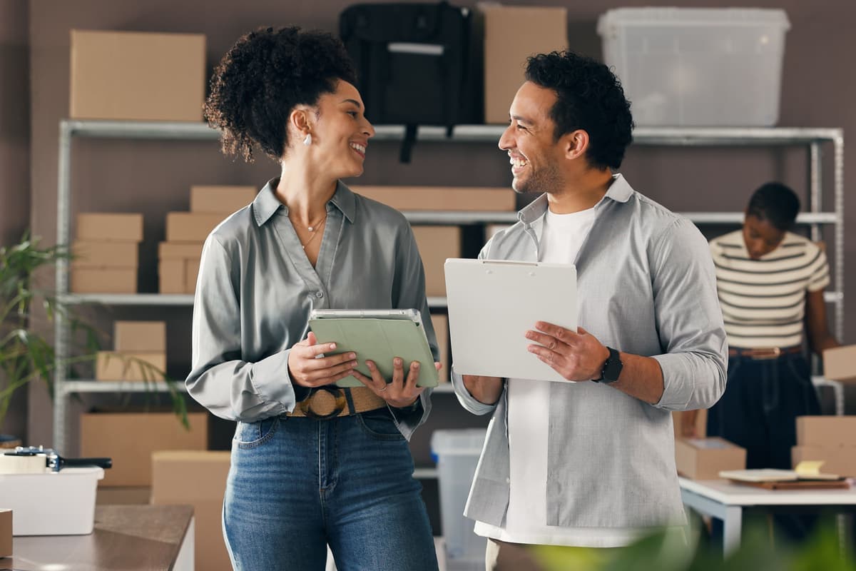 Two people smiling at each other while holding clipboards in a warehouse with boxes and a third person working in the background.
