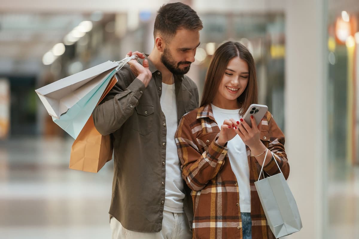 Smiling couple holding shopping bags and looking at a smartphone together while walking in a mall.