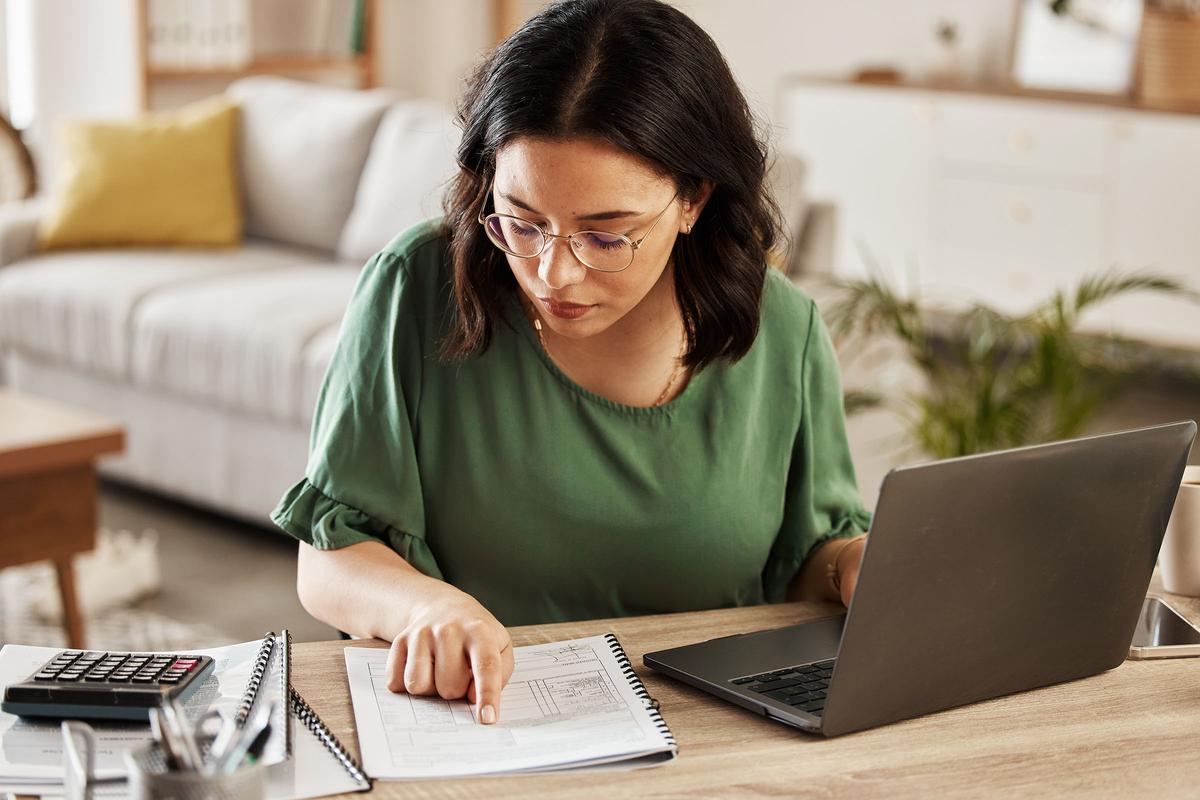 Woman in a green top working at a desk with a laptop, calculator, and documents, pointing to a page in a spiral notebook.