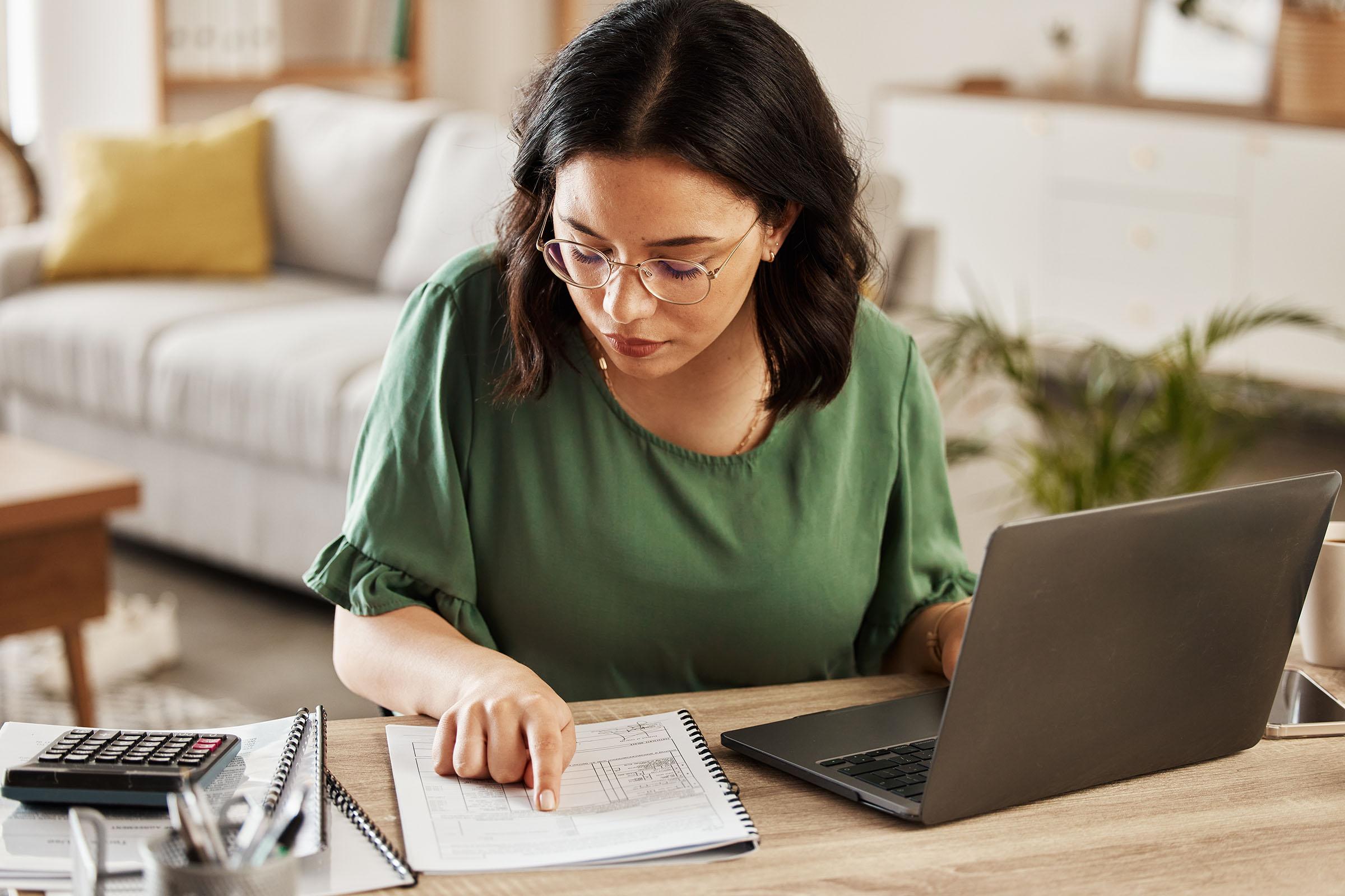 Woman in a green top working at a desk with a laptop, calculator, and documents, pointing to a page in a spiral notebook.