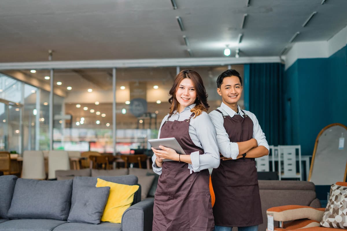 Two smiling employees in aprons stand in a furniture store, with sofas and seating items displayed around them.