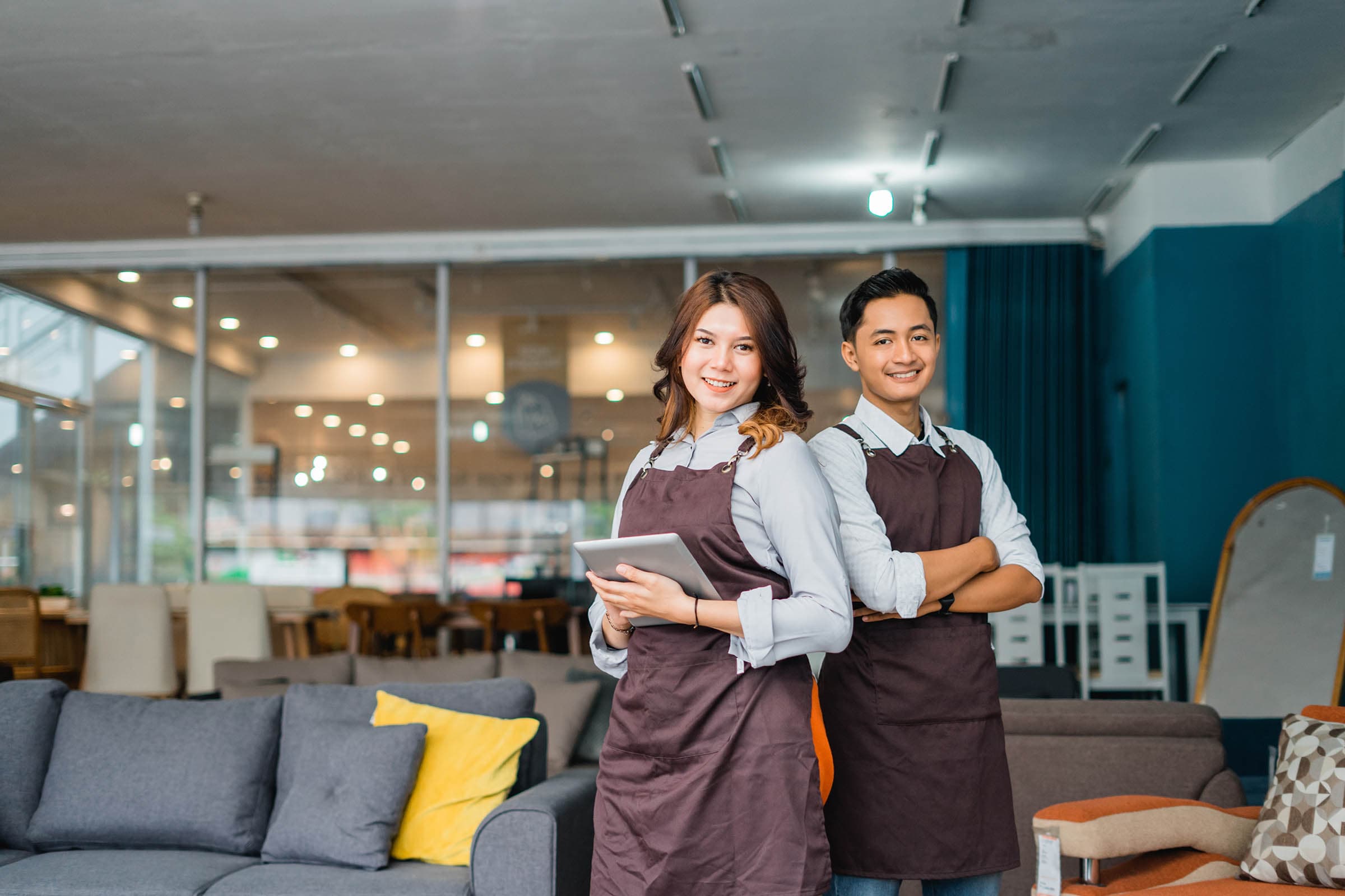 Two smiling employees in aprons stand in a furniture store, with sofas and seating items displayed around them.