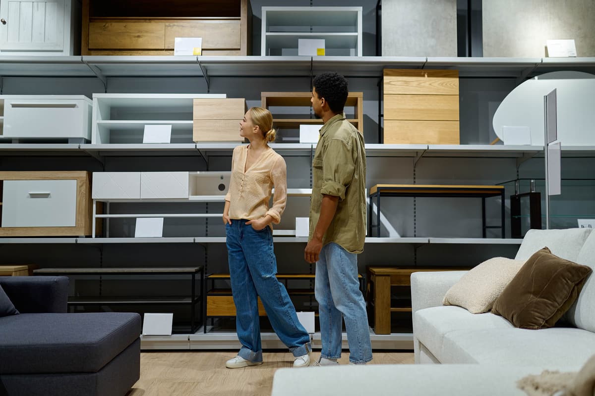 Two people in a furniture store browse wooden shelving units and cabinets on display shelves, with sofas in the foreground.