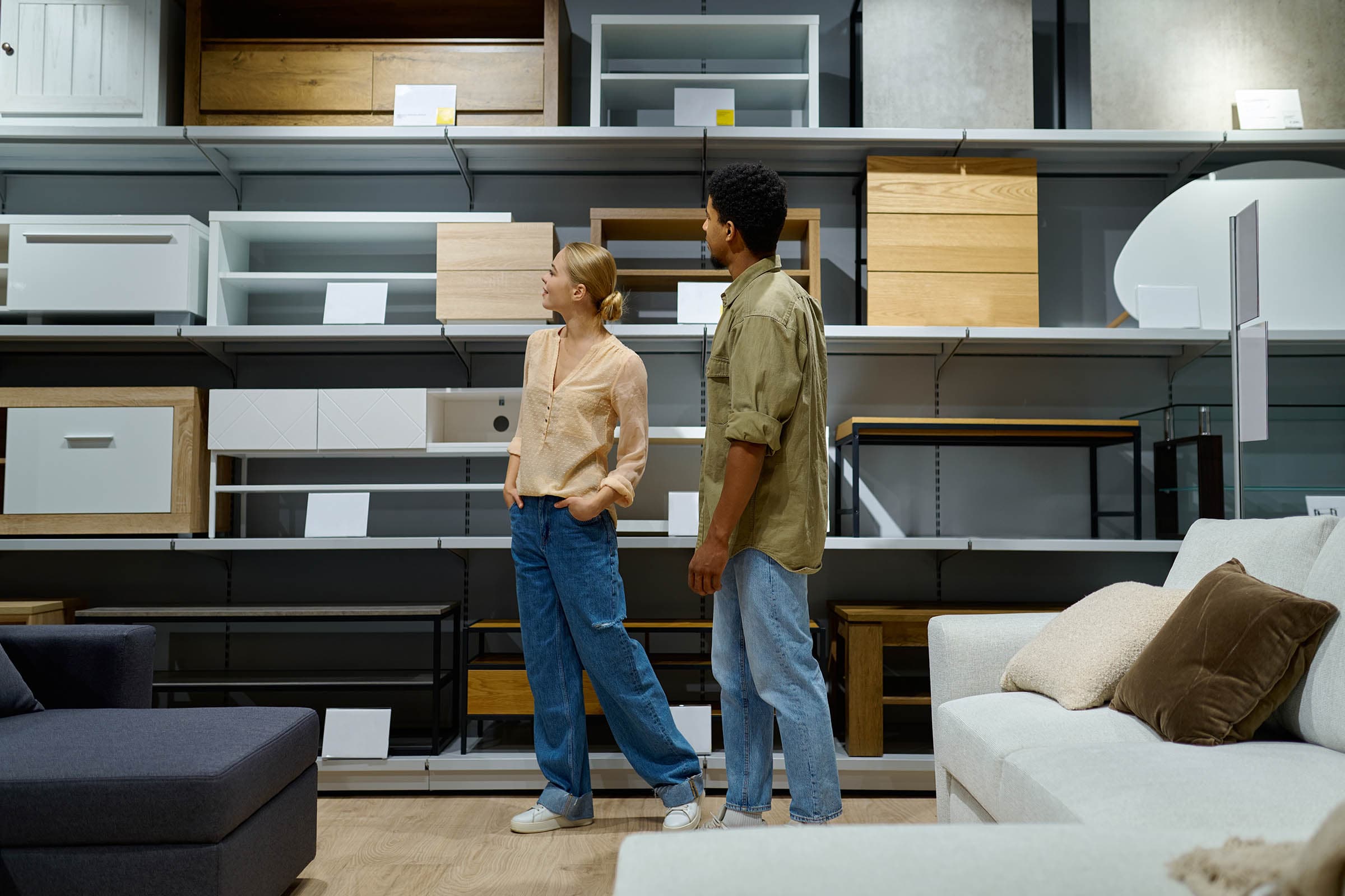 Two people in a furniture store browse wooden shelving units and cabinets on display shelves, with sofas in the foreground.