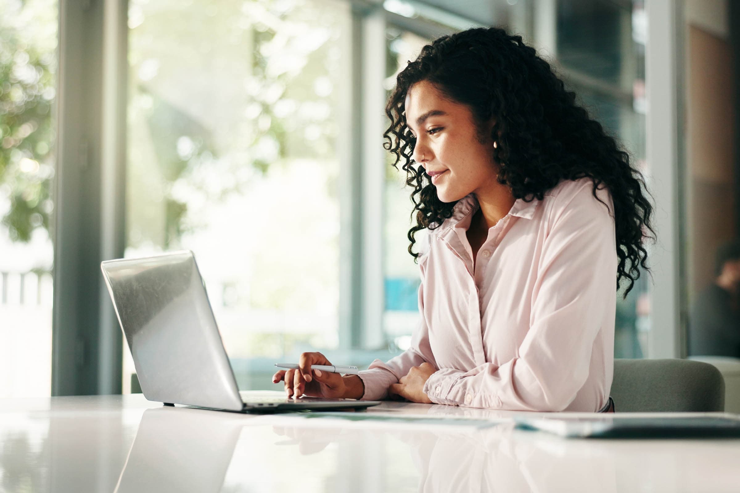 Woman with curly hair working on a laptop at a bright office desk, smiling and holding a pen.