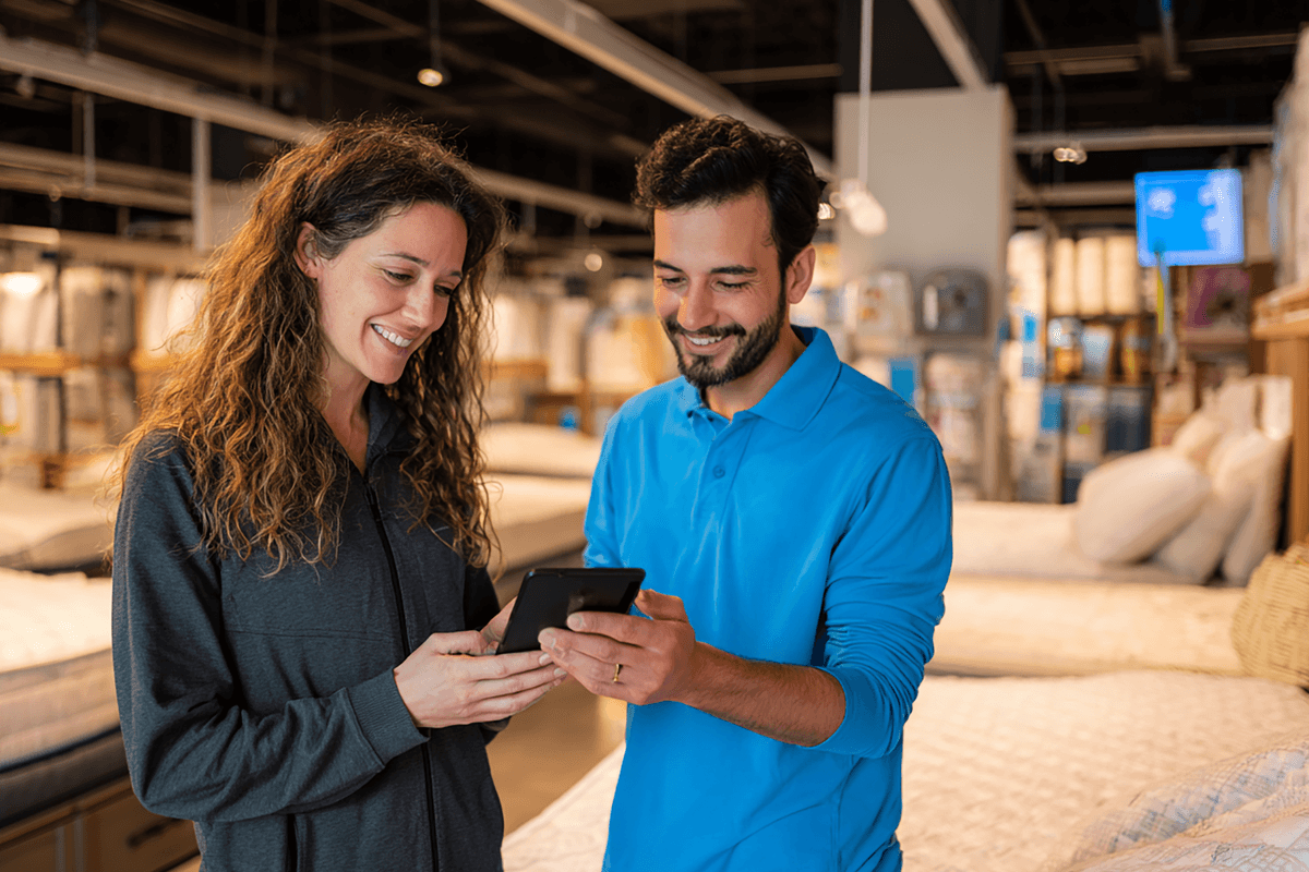 A woman and a man in a store smiling while looking at a phone. They are surrounded by mattresses and bedding products.