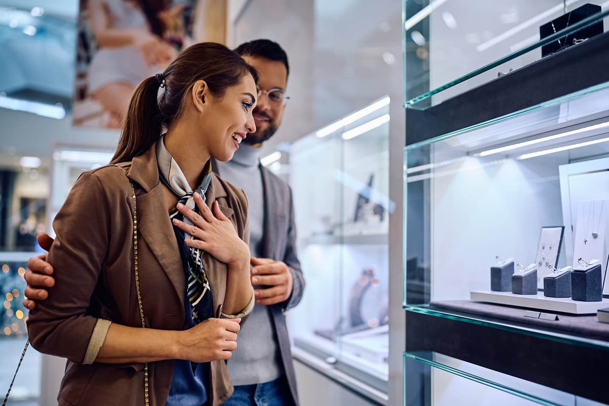 A couple joyfully window-shopping for jewelry in a store, with the woman looking excitedly at the display cases.