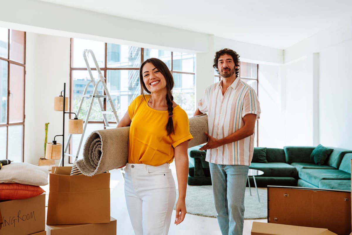 A smiling woman and a man carry a rolled-up rug in a bright living room with moving boxes and a green sofa.