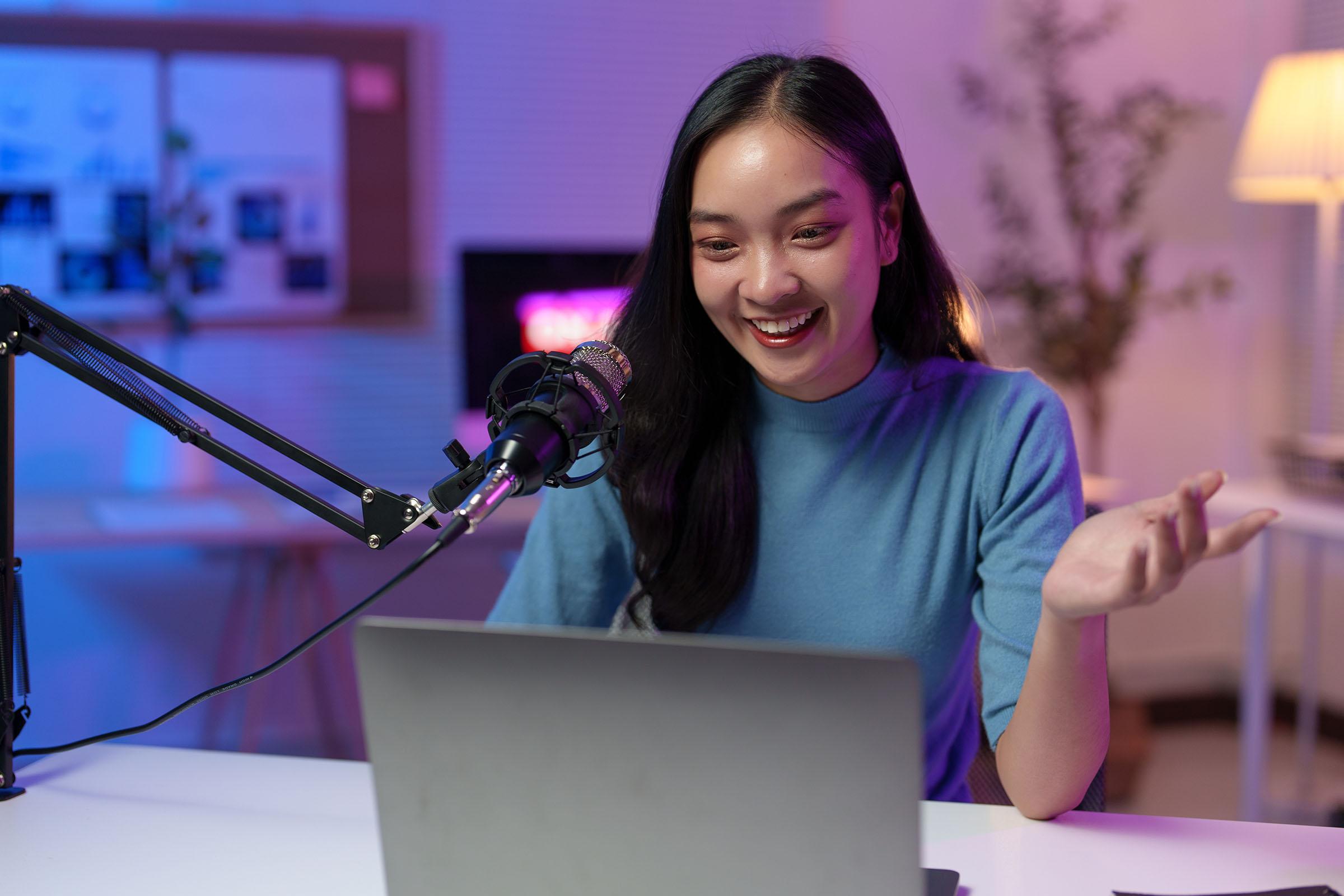 Smiling woman in a blue sweater speaking into a microphone during a livestream or podcast, seated at a desk with a laptop.