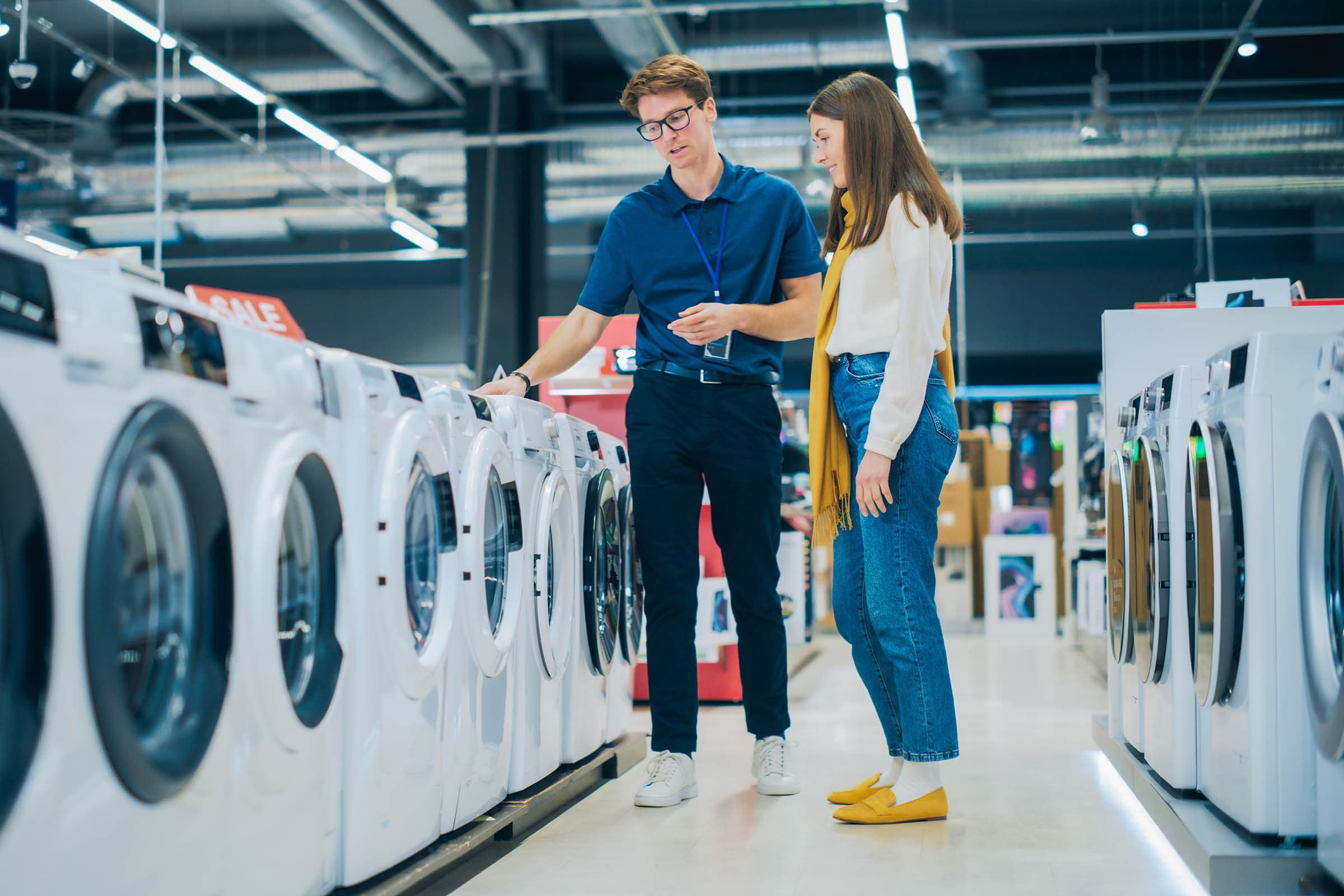 A salesperson shows a woman a row of washing machines in an appliance store, suggesting options with a sale sign in the background.