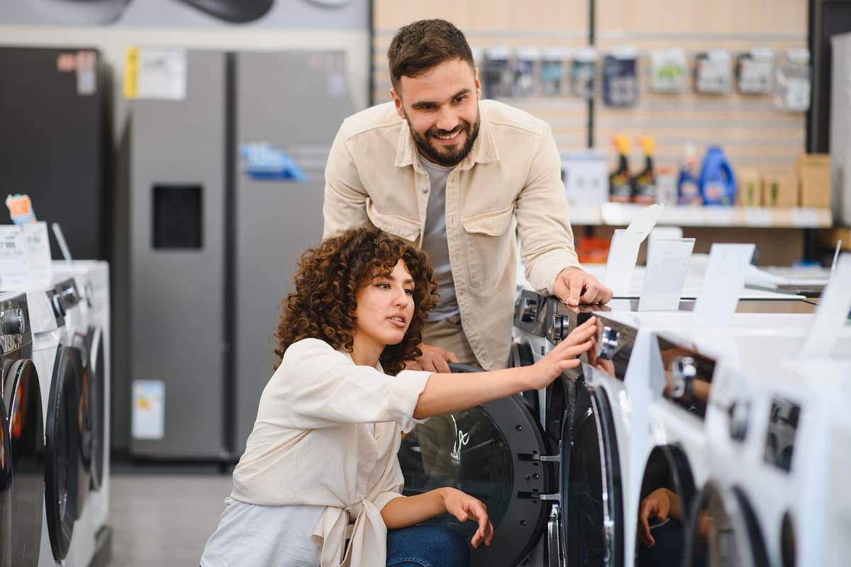 A couple in a store examines a row of washing machines. The man is standing, and the woman is kneeling, interacting with one machine.