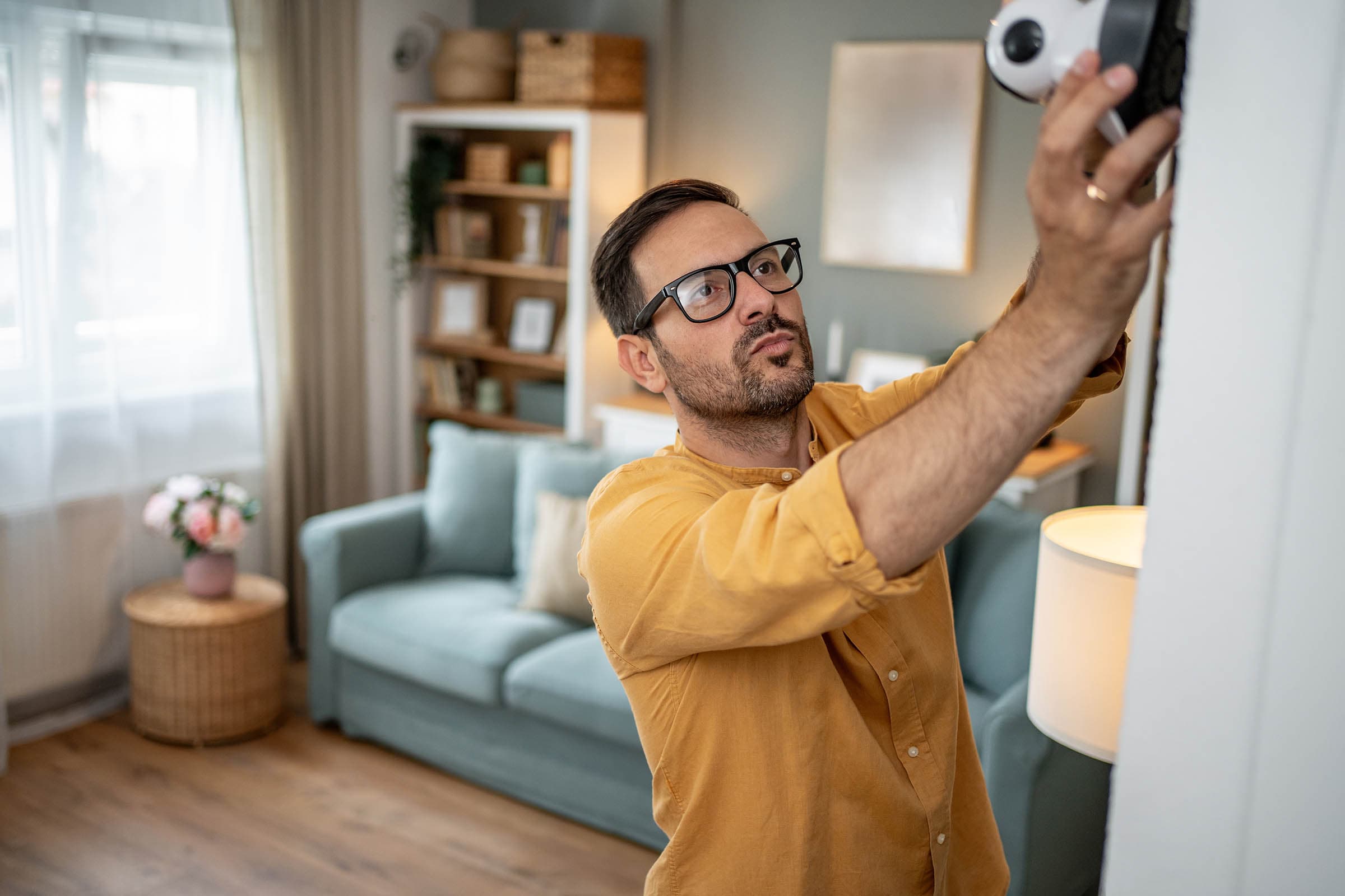 Man in glasses installs a home security camera on a wall in a cozy living room with a blue couch and bookshelf in the background.