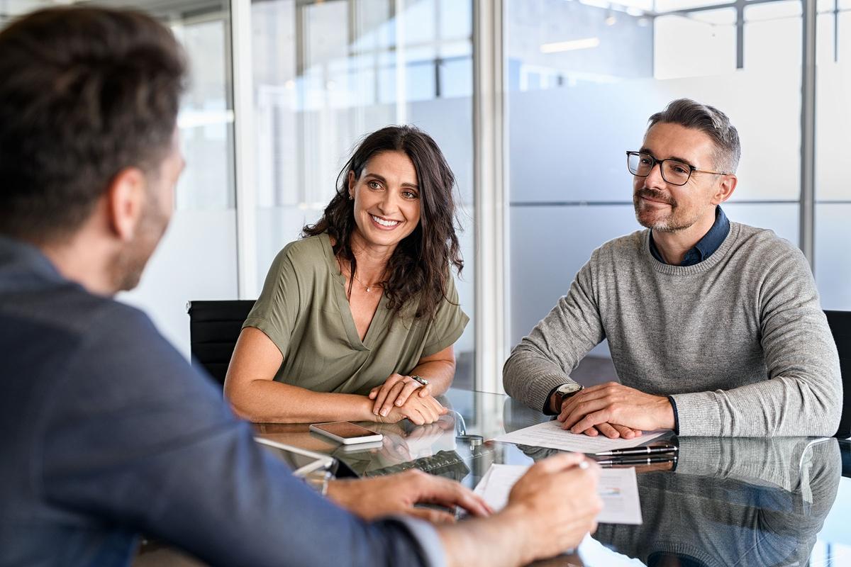 Smiling couple sitting at a glass table in an office, speaking with a professional holding documents.