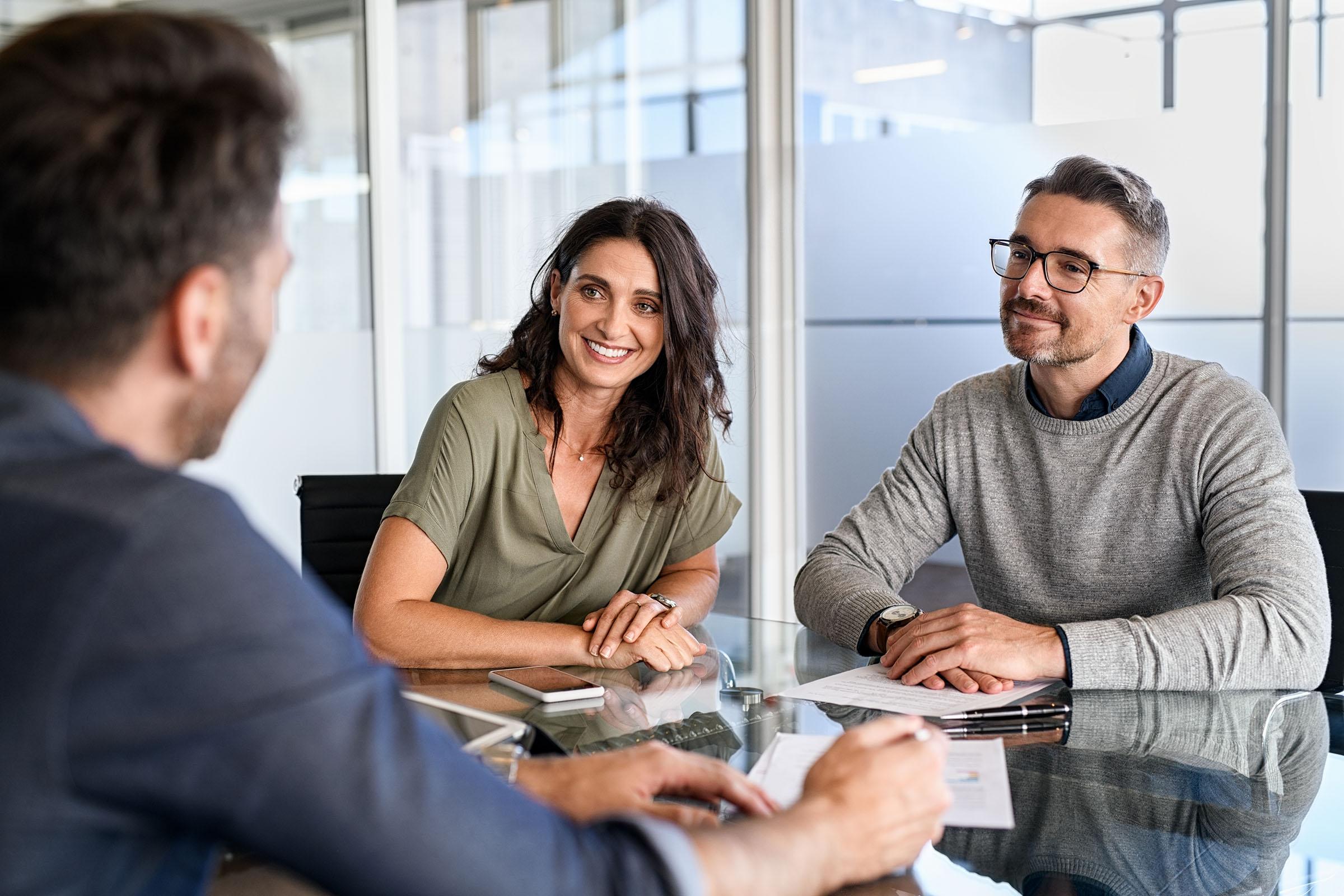 Smiling couple sitting at a glass table in an office, speaking with a professional holding documents.