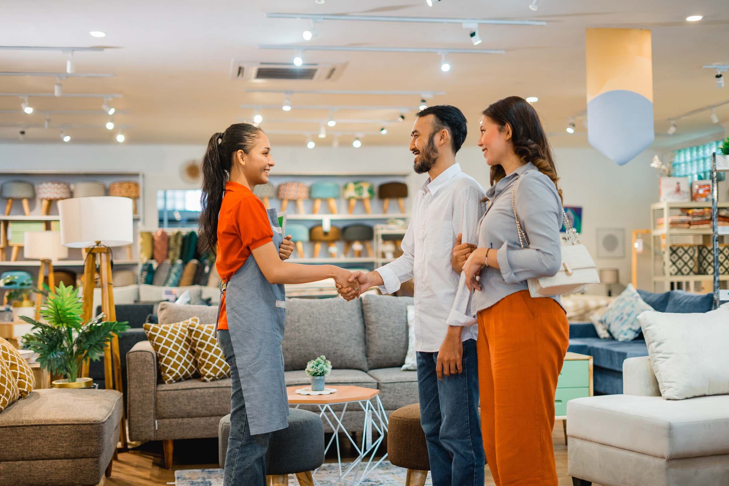 A store employee shakes hands with a couple in a furniture showroom filled with sofas and home decor items.