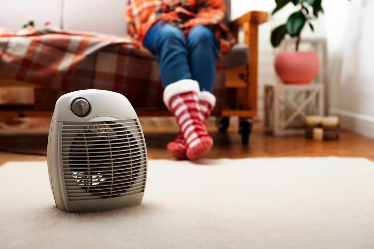 A small space heater is in the foreground, with a person in cozy socks sitting on a sofa in the background.