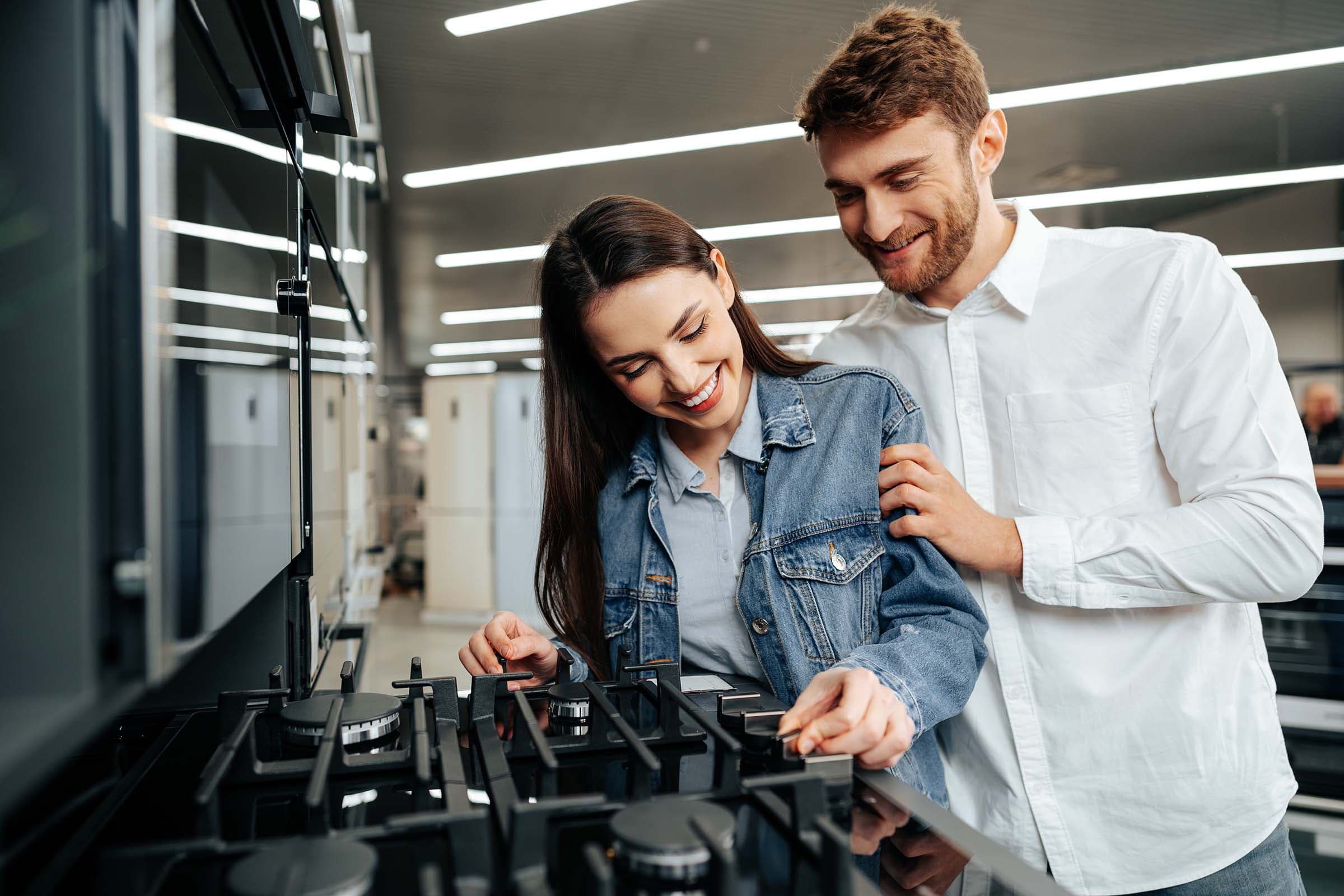 A smiling woman adjusts knobs on a stove in a showroom, while a man stands beside her with a hand on her shoulder.