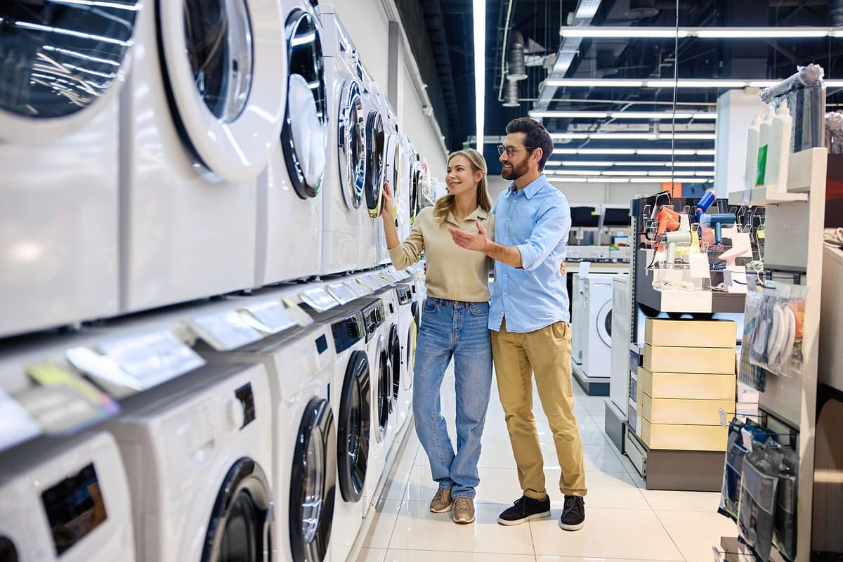 A couple shopping for washing machines in an appliance store, smiling and examining the options on display.