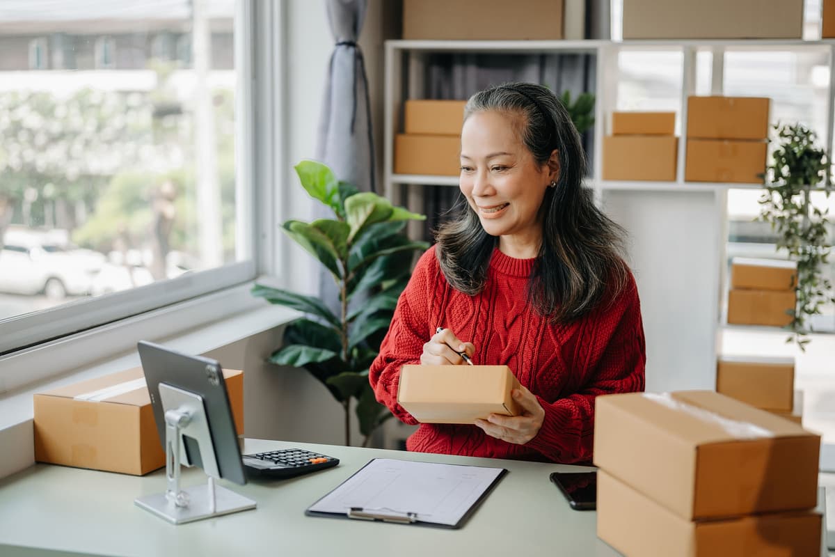 A woman in a red sweater writes on a package at a desk, surrounded by boxes, with a tablet, clipboard, and calculator nearby.
