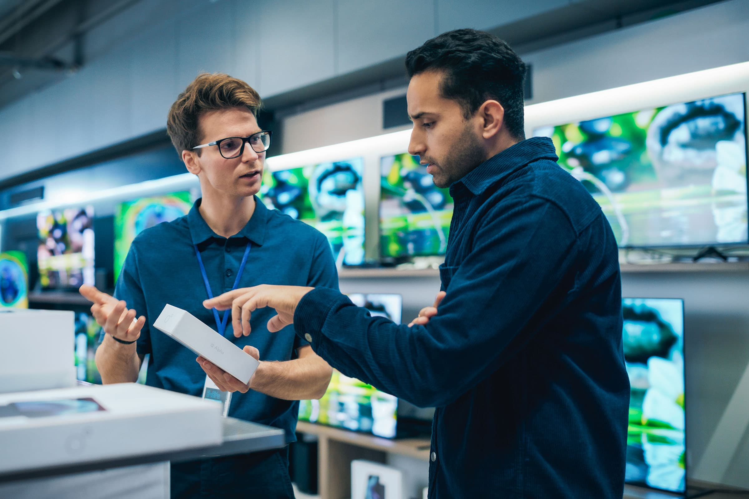 A store employee is explaining a product to a customer in an electronics shop, surrounded by flat-screen TVs displaying colorful images.
