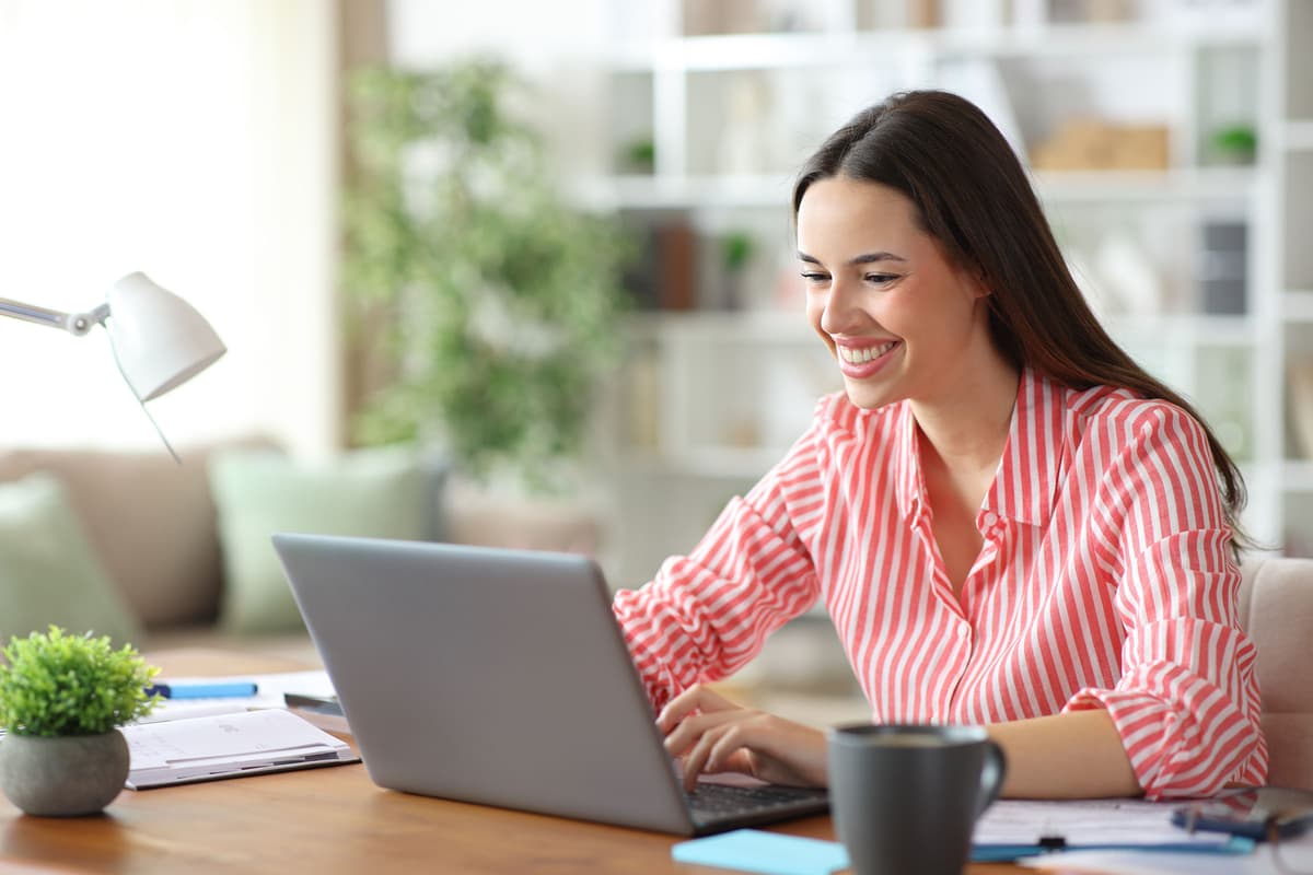 Smiling woman in a pink striped shirt working on a laptop at a desk, with a notebook, cup, lamp, and potted plant. Bright, airy background.