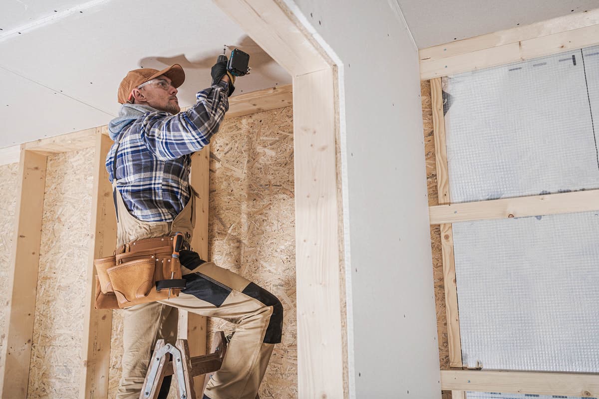 Carpenter on ladder installing drywall in a wooden-framed interior, wearing a plaid shirt, cap, and tool belt, using a drill overhead.