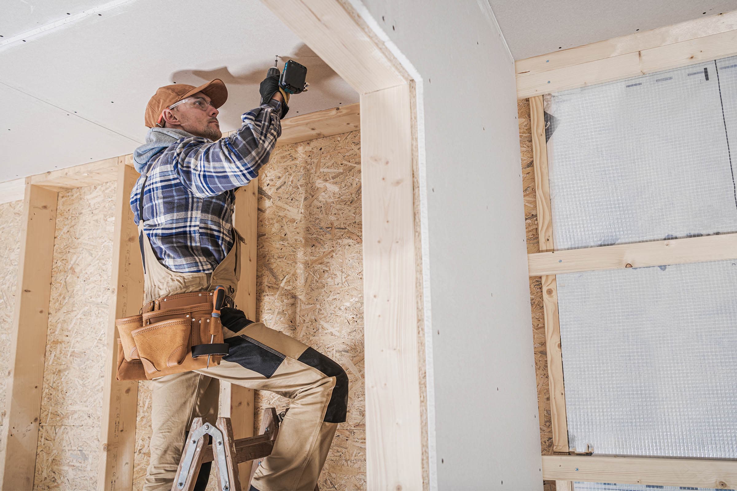 Carpenter on ladder installing drywall in a wooden-framed interior, wearing a plaid shirt, cap, and tool belt, using a drill overhead.