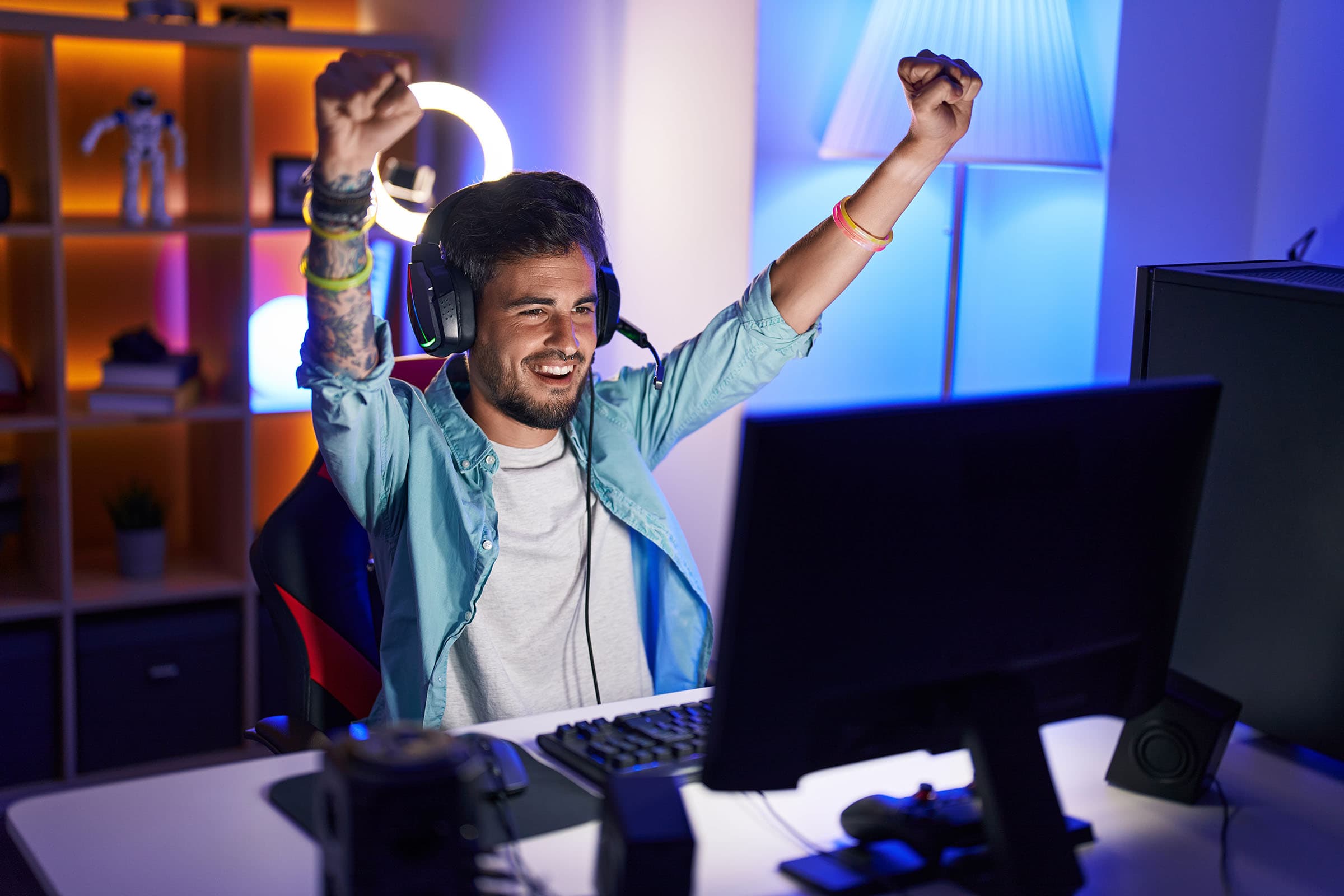 A man wearing headphones celebrates in front of a computer screen, with colorful backlighting in a gaming setup.