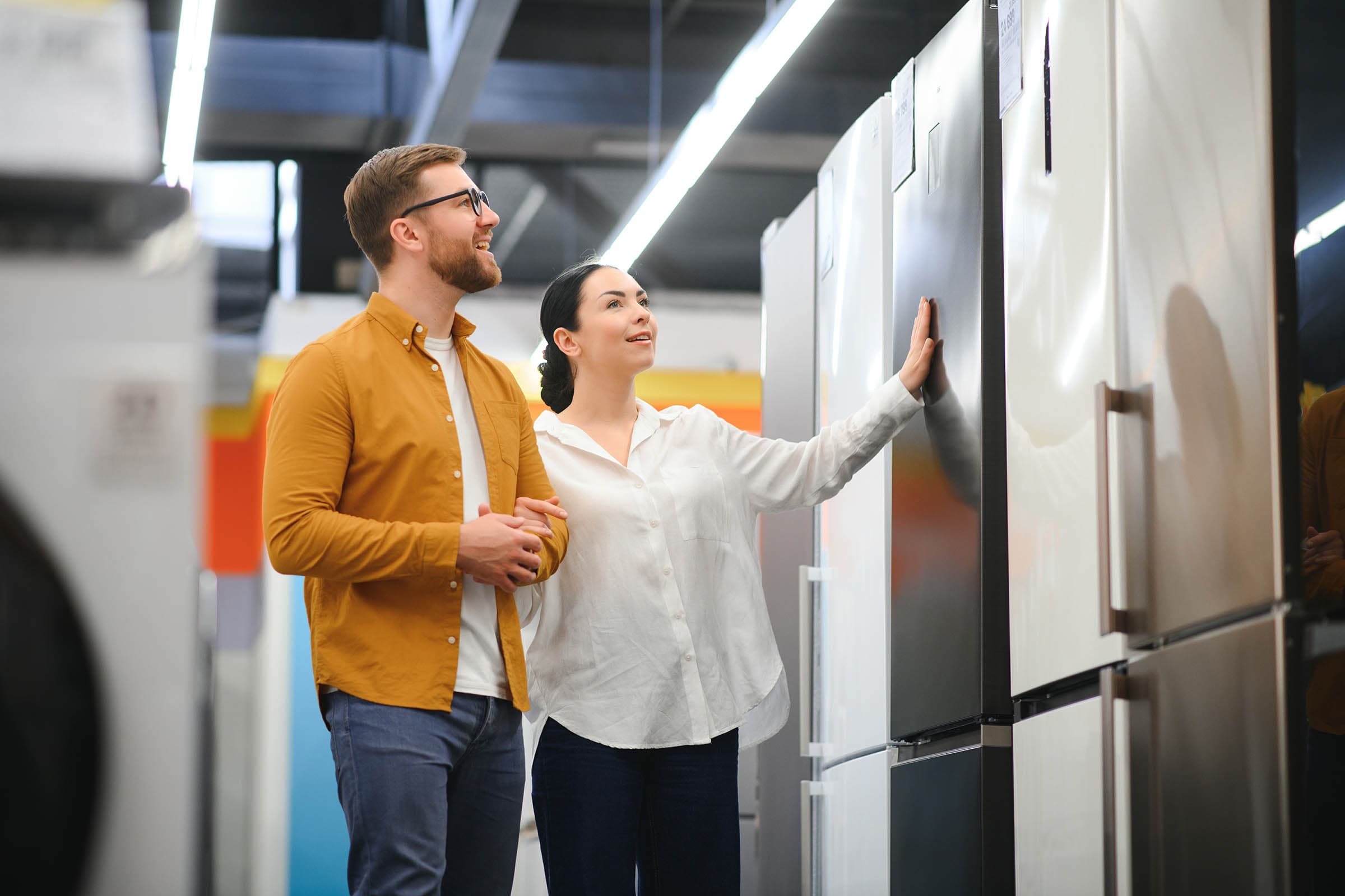 A couple examines refrigerators in a brightly lit appliance store, with colorful shelves in the background.