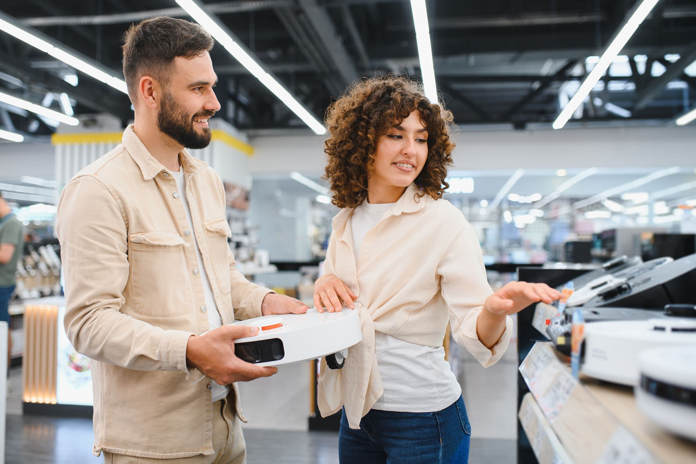 A man and woman in a store examine robotic vacuum cleaners. The man holds one, and the woman points at models on display.