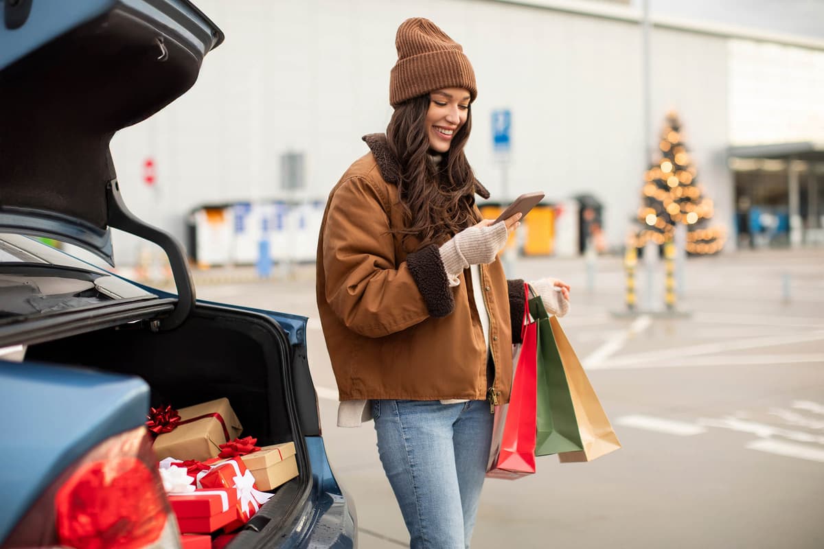 Woman in winter clothing checks her phone, holding shopping bags by an open car trunk with gift boxes. Decorated tree in the background.