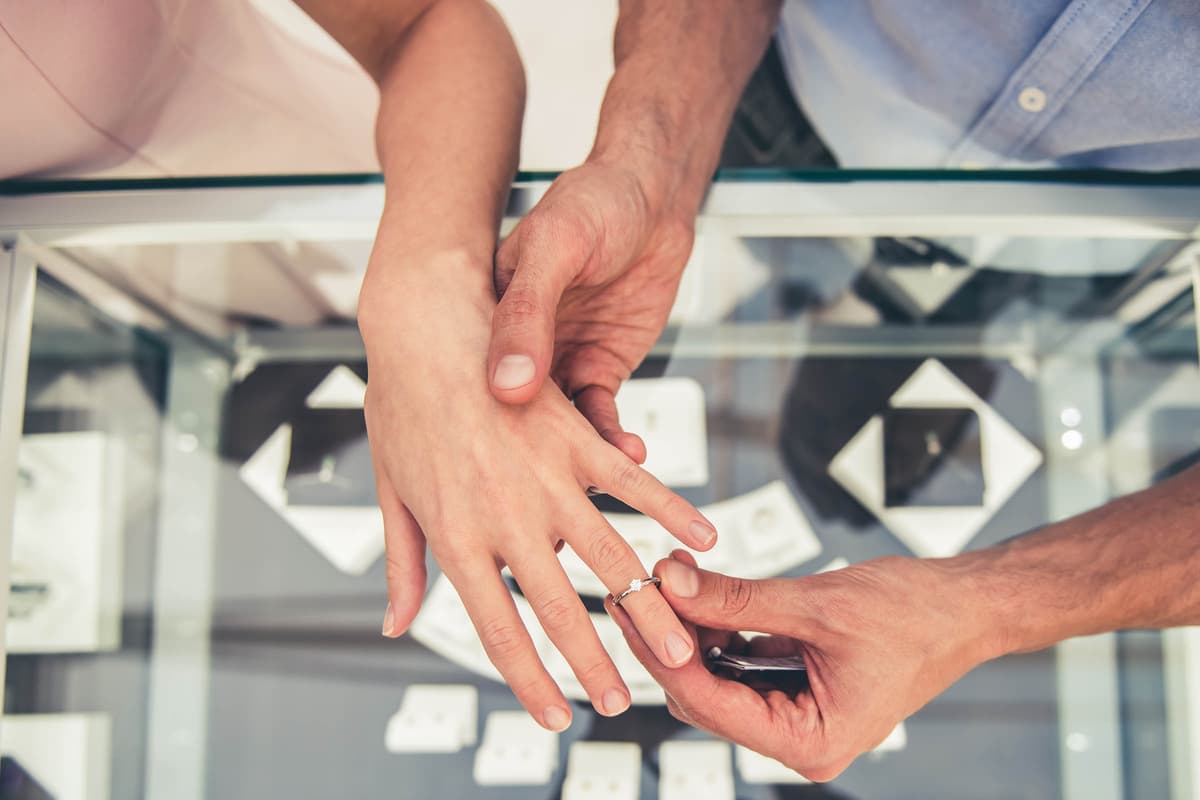 Close-up of a person placing a diamond ring on another's finger, with a jewelry store display in the background.
