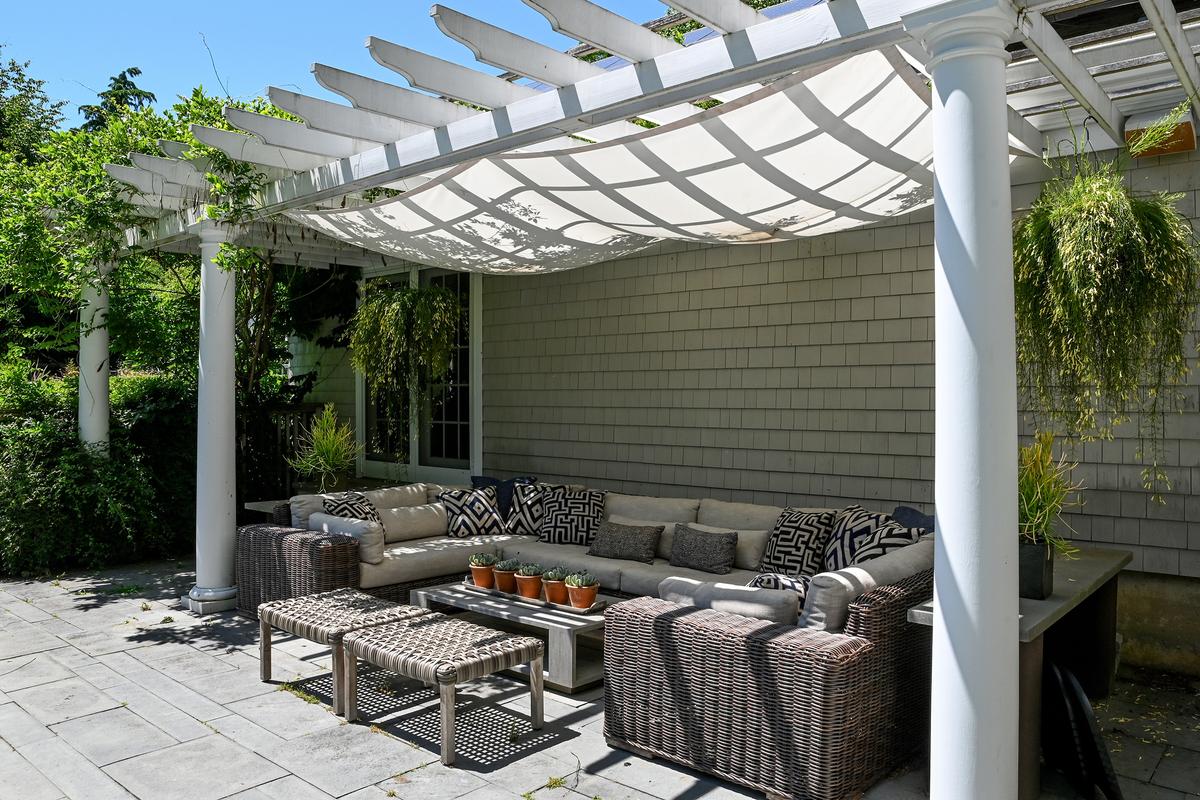 Shaded patio beneath white pergola with wicker sectional, beige cushions, patterned pillows, low table, and potted succulents.