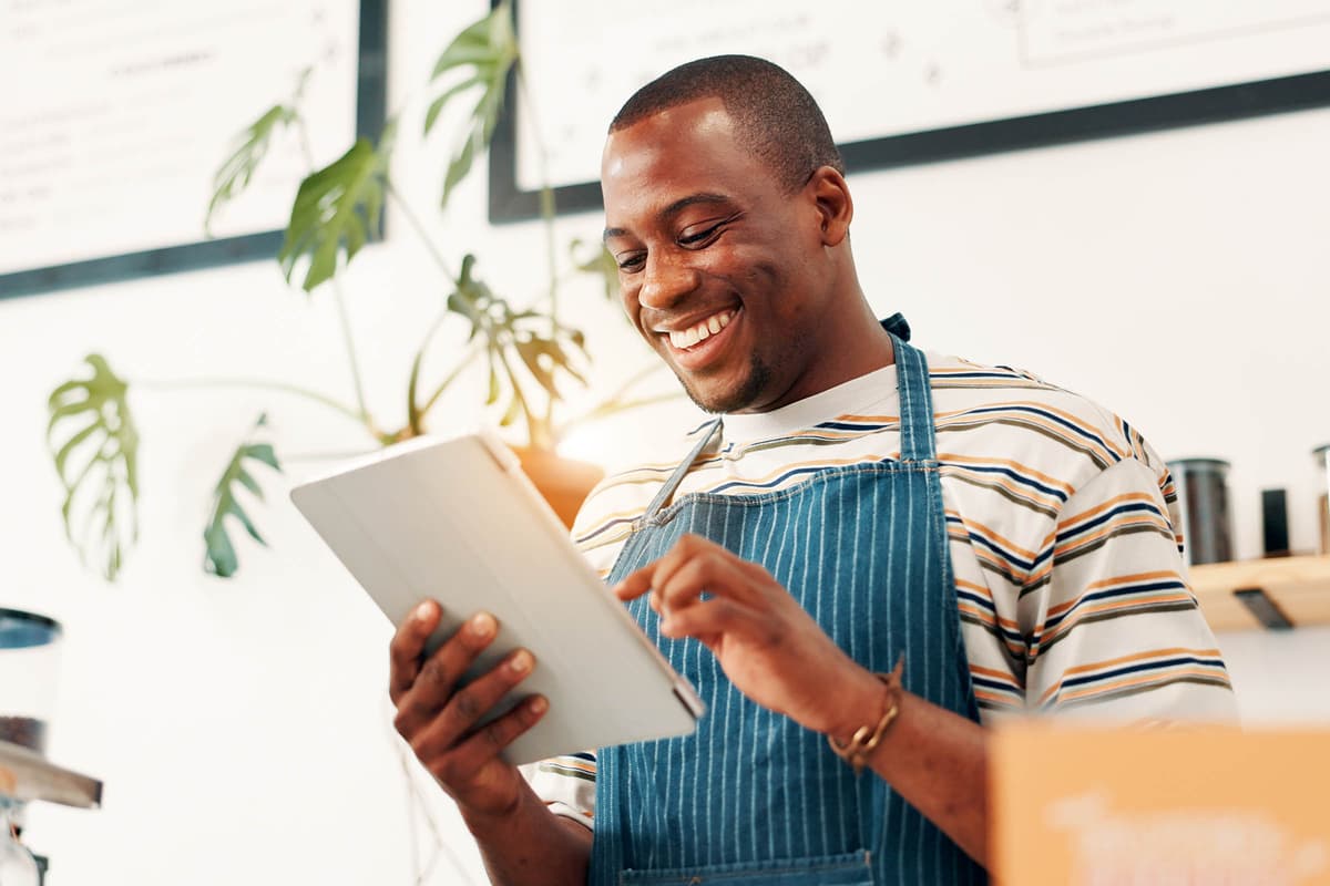 Smiling person in a striped shirt and apron uses a tablet in a bright room with plants and shelves.