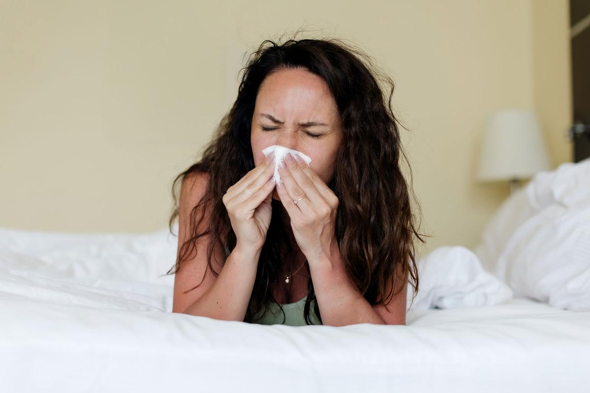 Woman lying in bed with eyes closed, sneezing or blowing her nose into a tissue, appearing to be unwell.