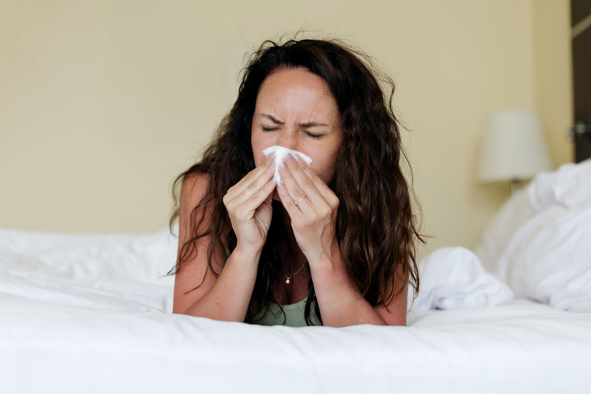 Woman lying in bed with eyes closed, sneezing or blowing her nose into a tissue, appearing to be unwell.