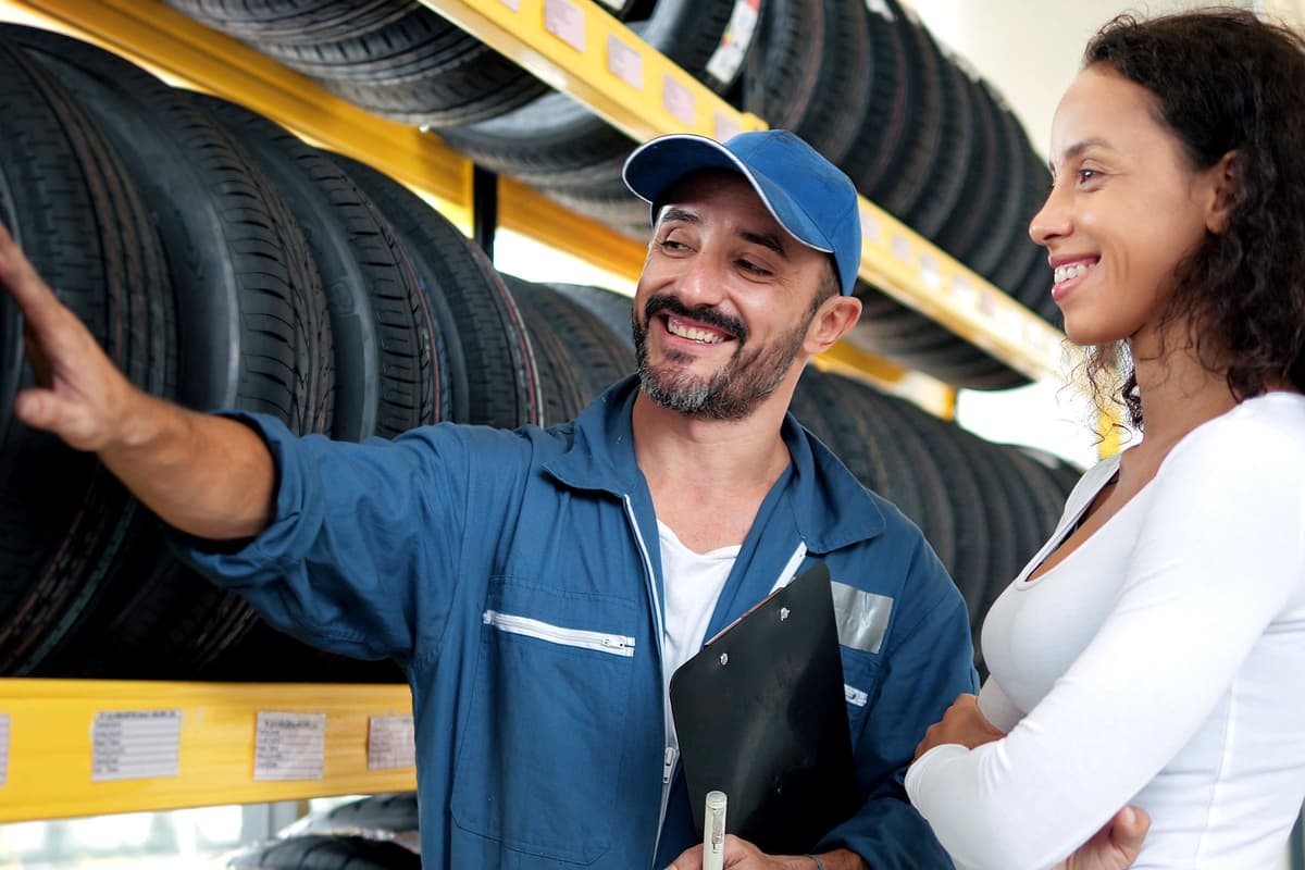 Smiling mechanic in a blue uniform and cap showing tire options to a customer in a tire shop.