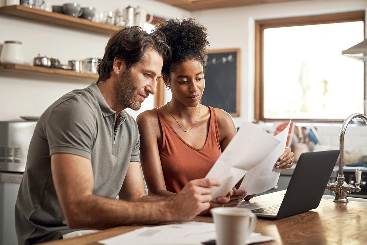 A couple sits at a kitchen table reviewing documents together, with a laptop open in front of them and papers spread out on the table.