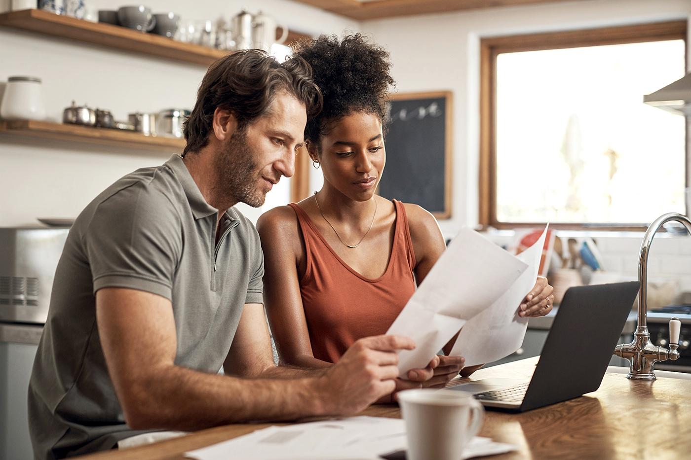 A couple sits at a kitchen table reviewing documents together, with a laptop open in front of them and papers spread out on the table.